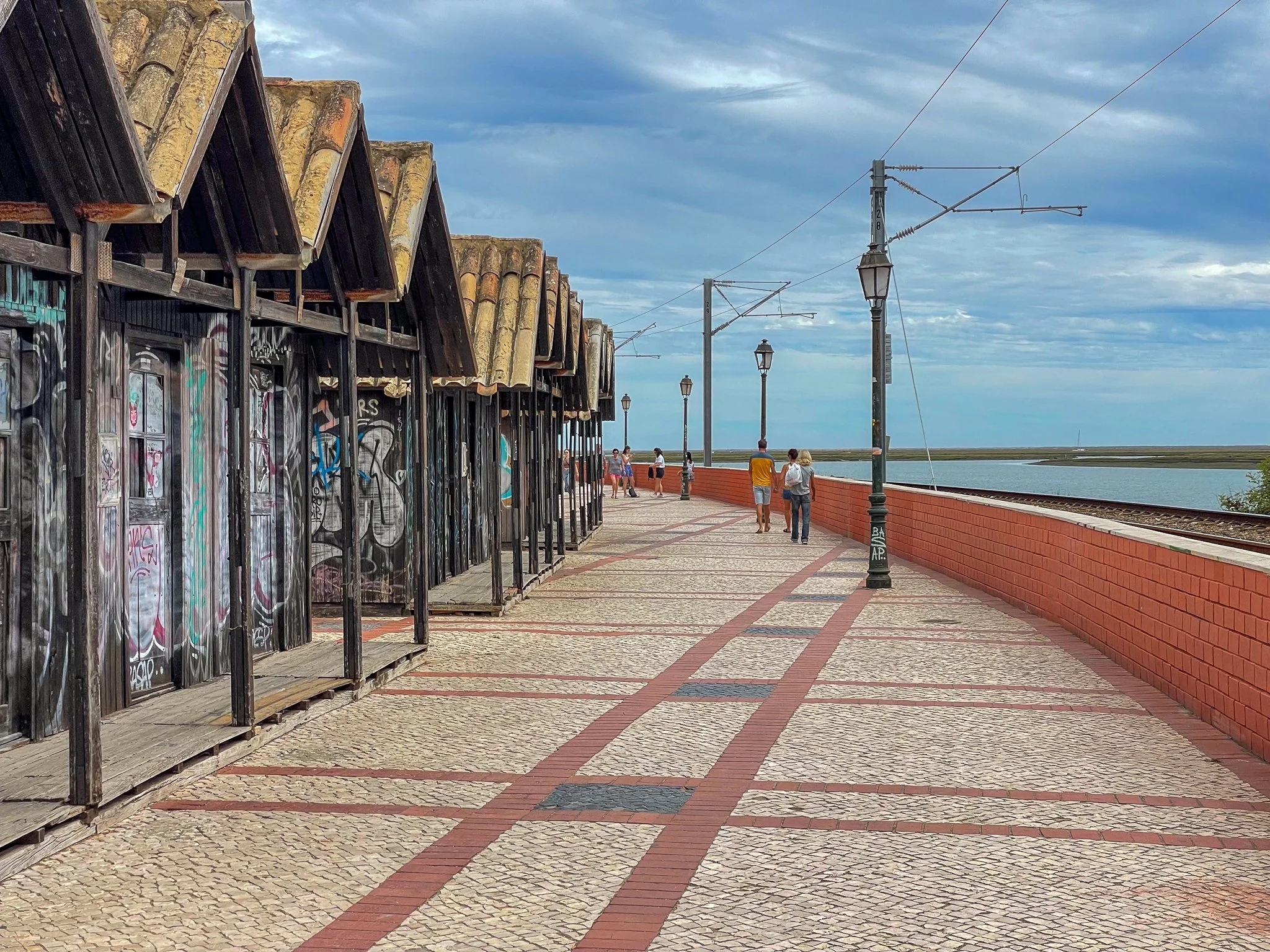 A film and photo location in Portugal from walnut films, A seaside promenade with a patterned stone walkway, a row of old, graffiti-covered shuttered shops on the left, decorative street lamps, and a red brick railing. Several people are walking, 
