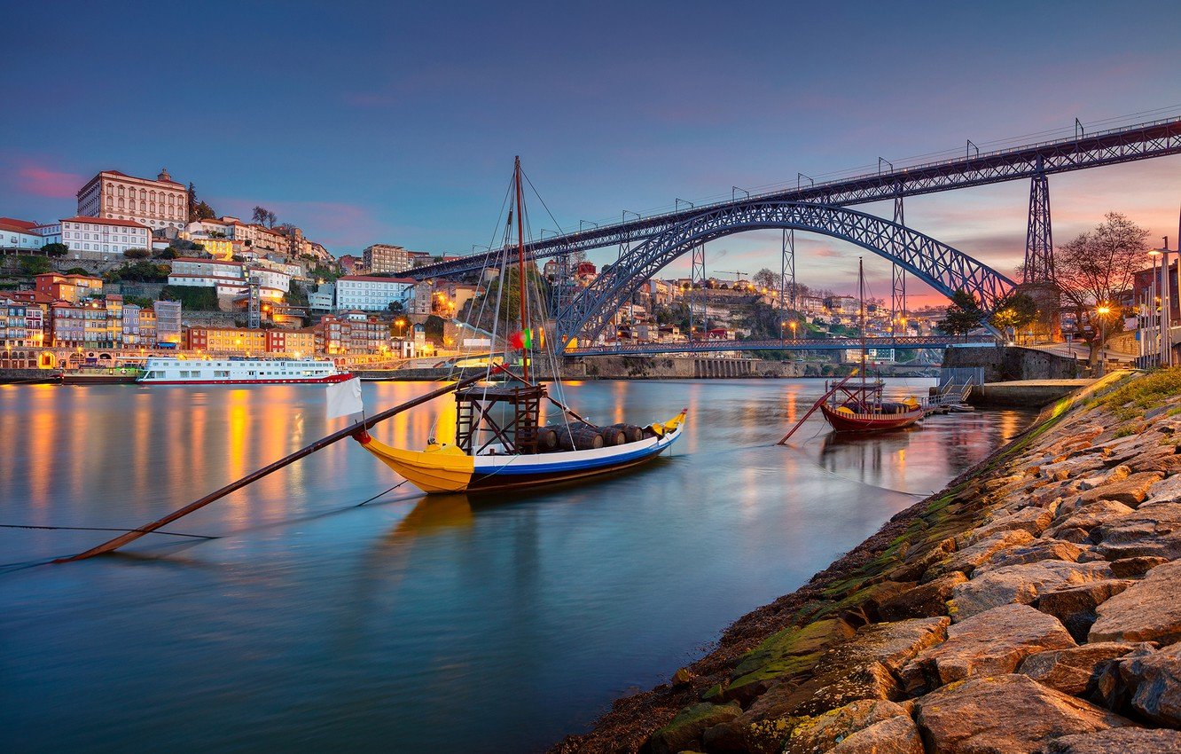 A film and photo location in Portugal from walnut films, A scenic view of Porto, Portugal at sunset, with colorful houses on a hillside, a bridge spanning the river, and boats docked along the waterfront.