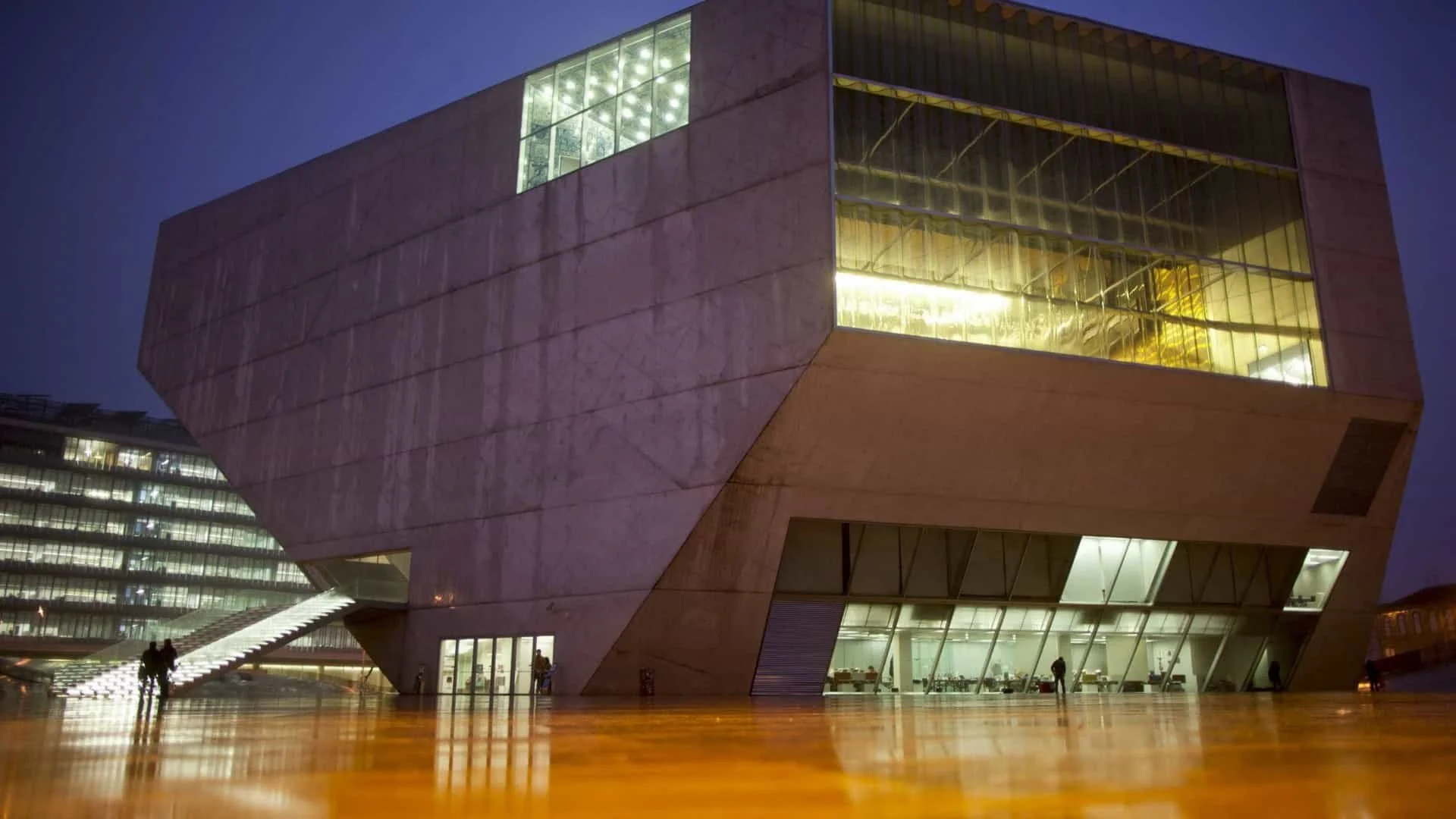 A film and photo location in Portugal from walnut films, Modern architectural building with large glass windows, illuminated from within, at night. Some people are visible outside on a reflective wooden plaza.
