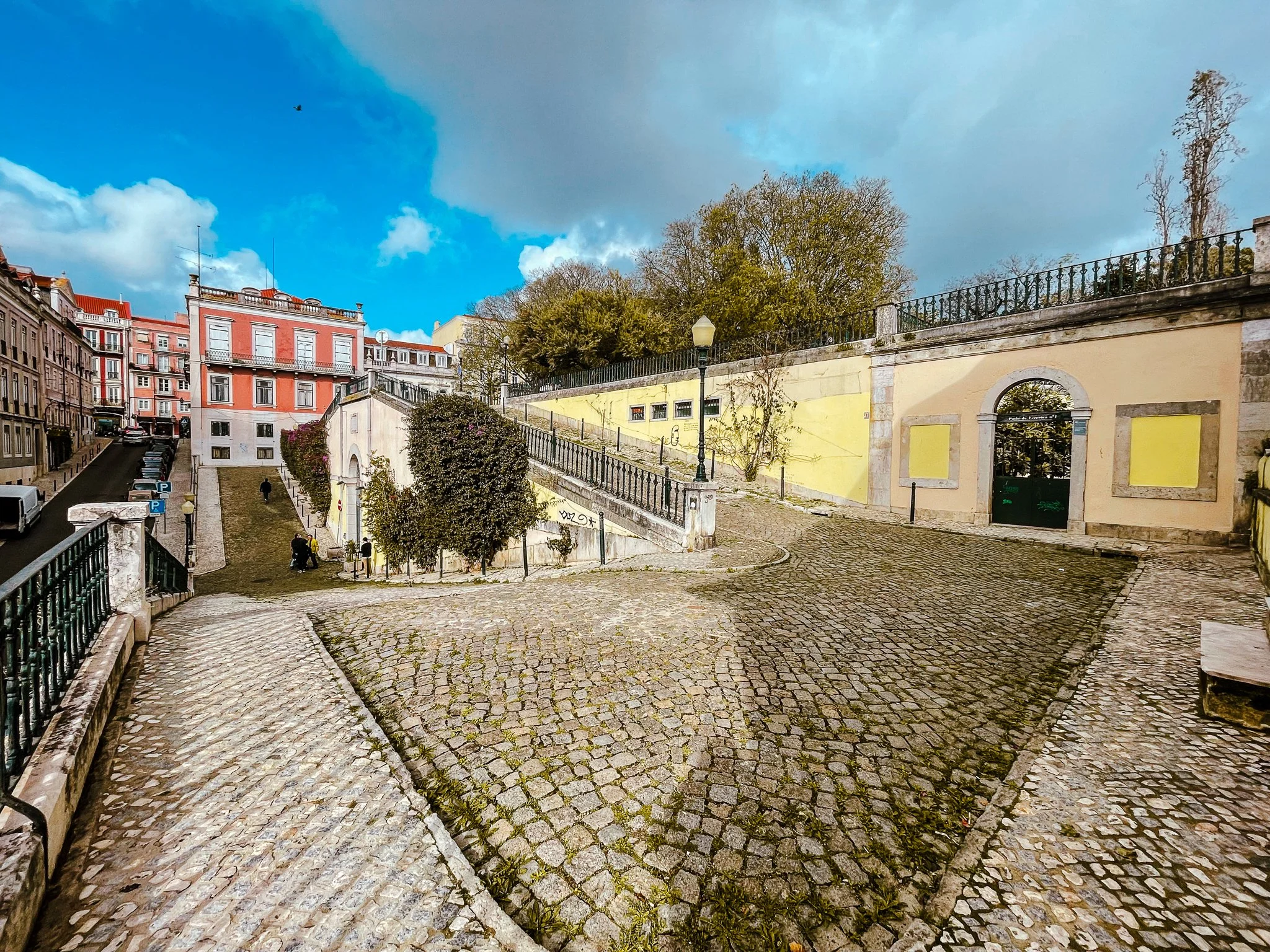 A film and photo location in Portugal from walnut films, Cobblestone street with stairs, black wrought iron fence, trees, historic buildings, and a partly cloudy sky.