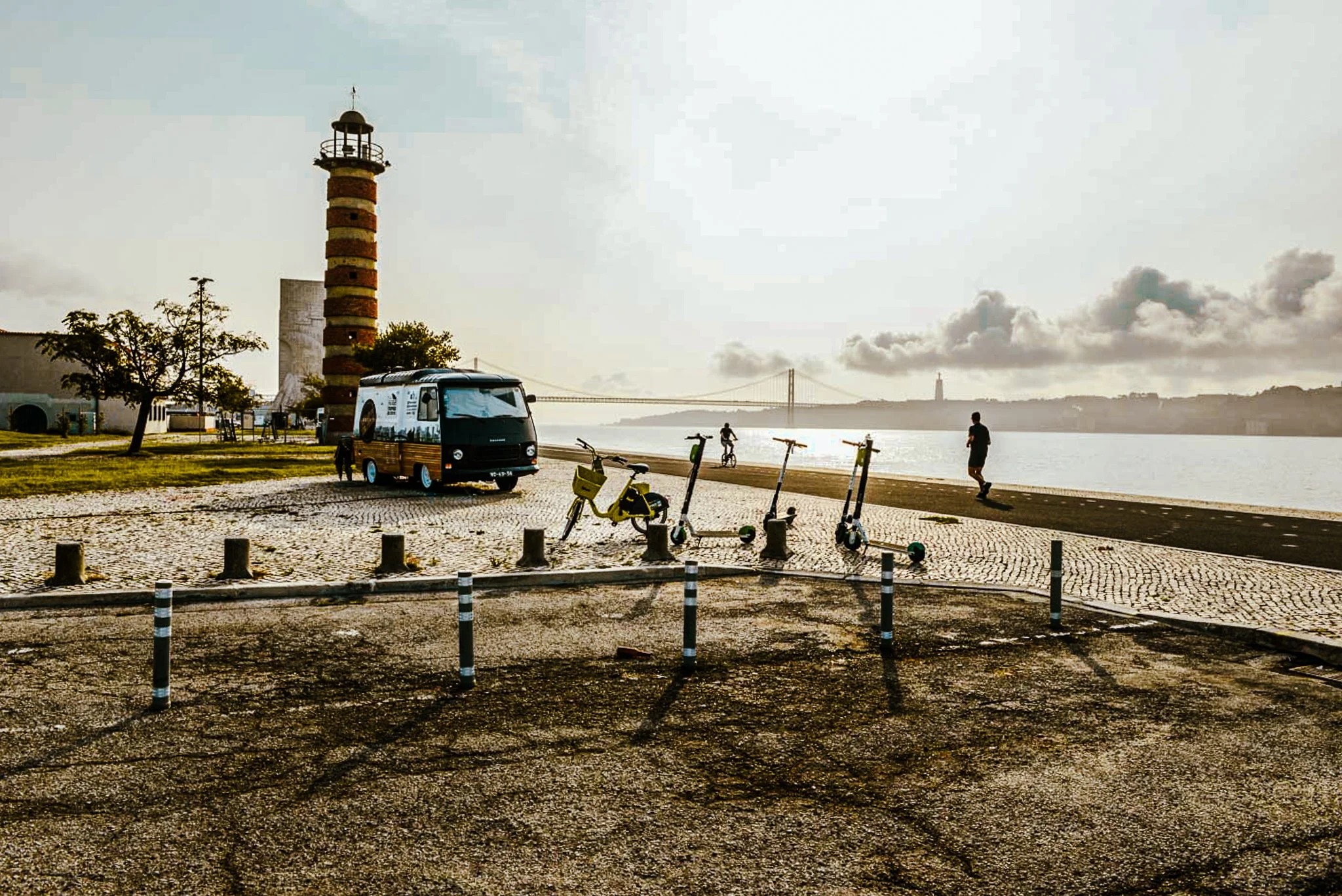 A film and photo location in Portugal from walnut films, A scenic waterfront scene during sunset with a lighthouse on the left, several people walking or riding bikes along the path, a parked bus, and scooters lined along the sidewalk. 