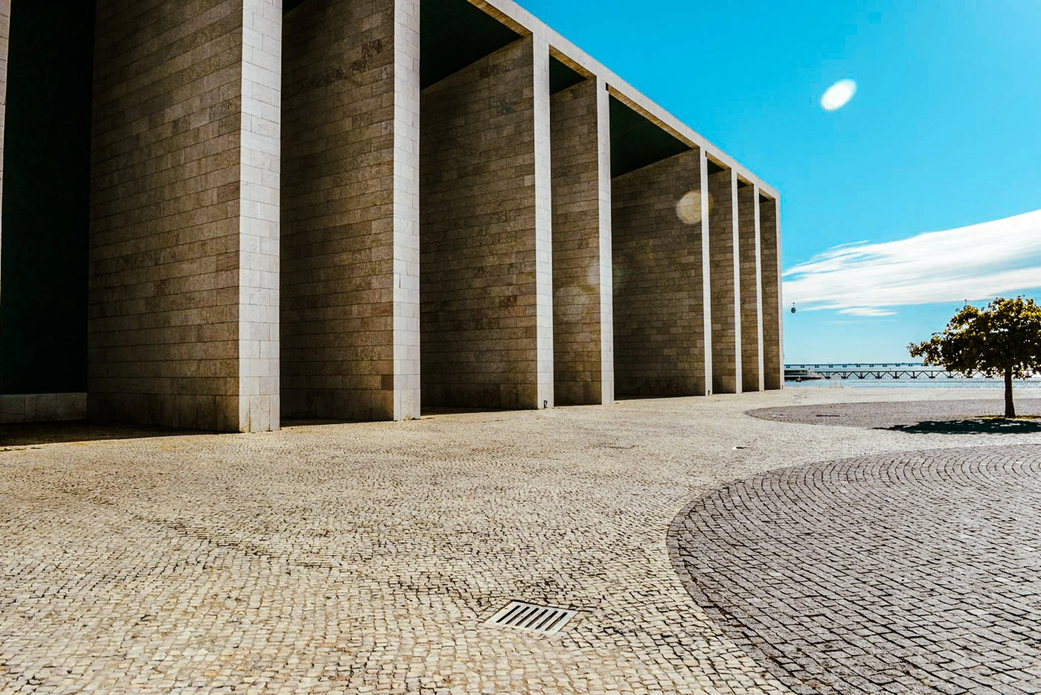 A film and photo location in Portugal from walnut films, Modern building with tall, brick walls and white vertical supports along a paved open plaza. A tree is on the right, and the sky is partly cloudy with a bright sun.