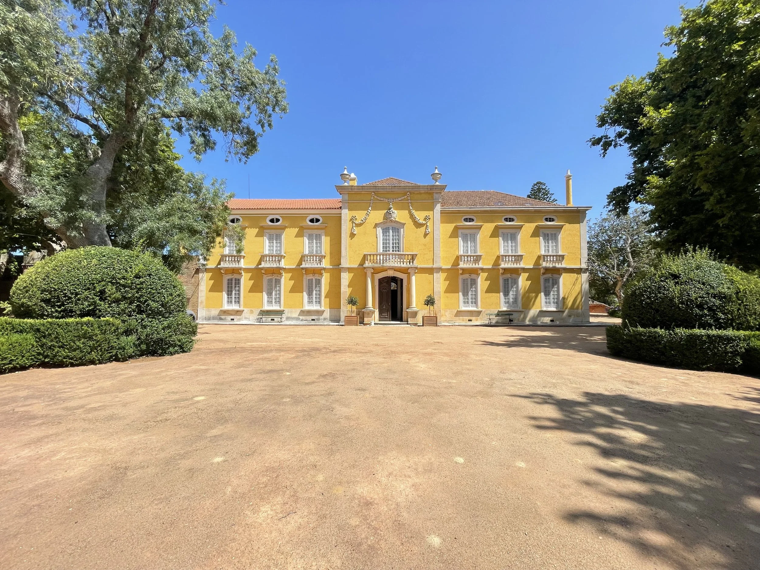 A film and photo location in Portugal from walnut films, A large yellow mansion with colonial architectural features, surrounded by trees and bushes, under a blue sky.
