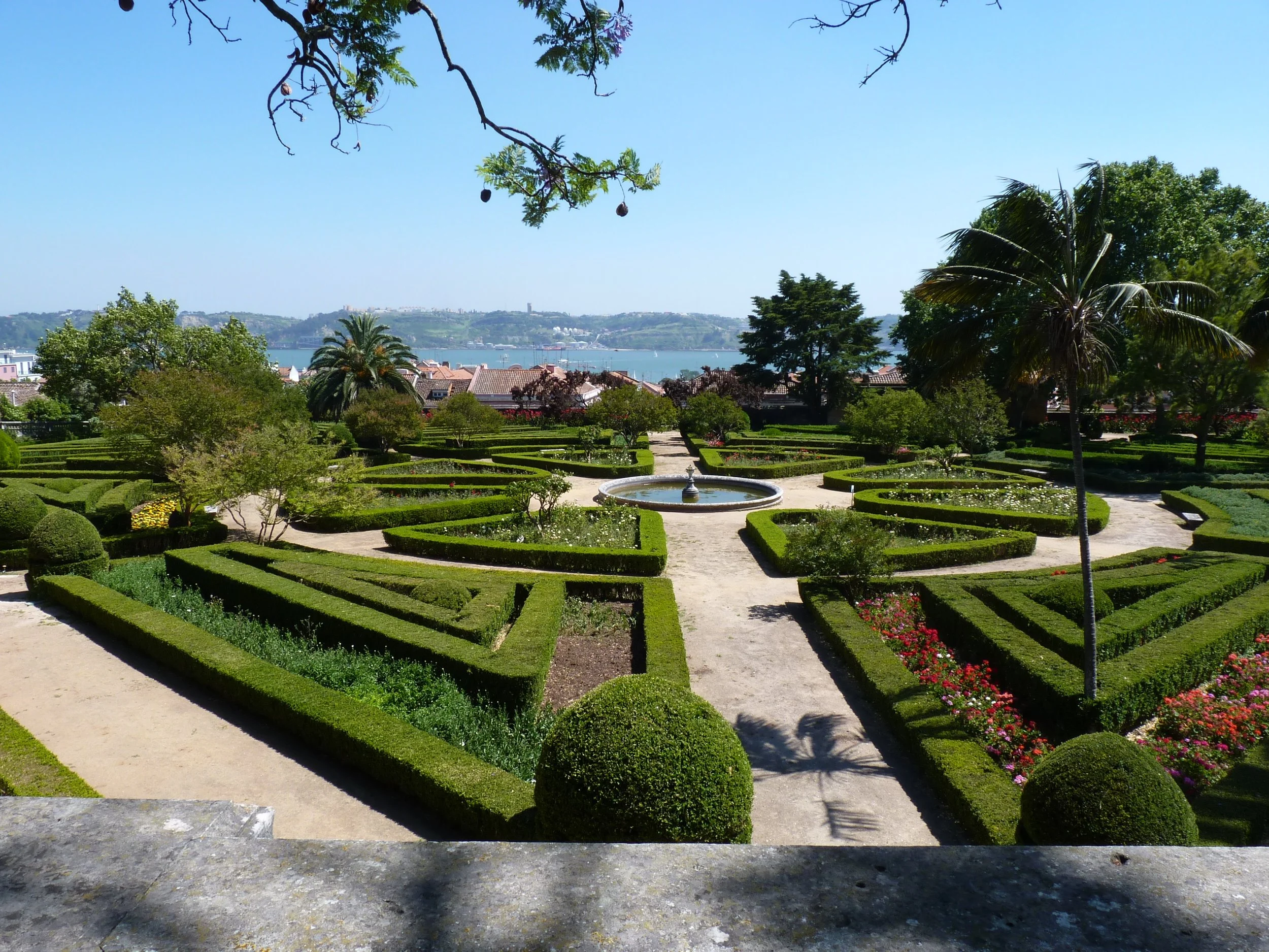 A film and photo location in Portugal from walnut films, View of a formal garden with trimmed hedges, flower beds, a fountain, and paths, with water and hills in the background under a clear sky.