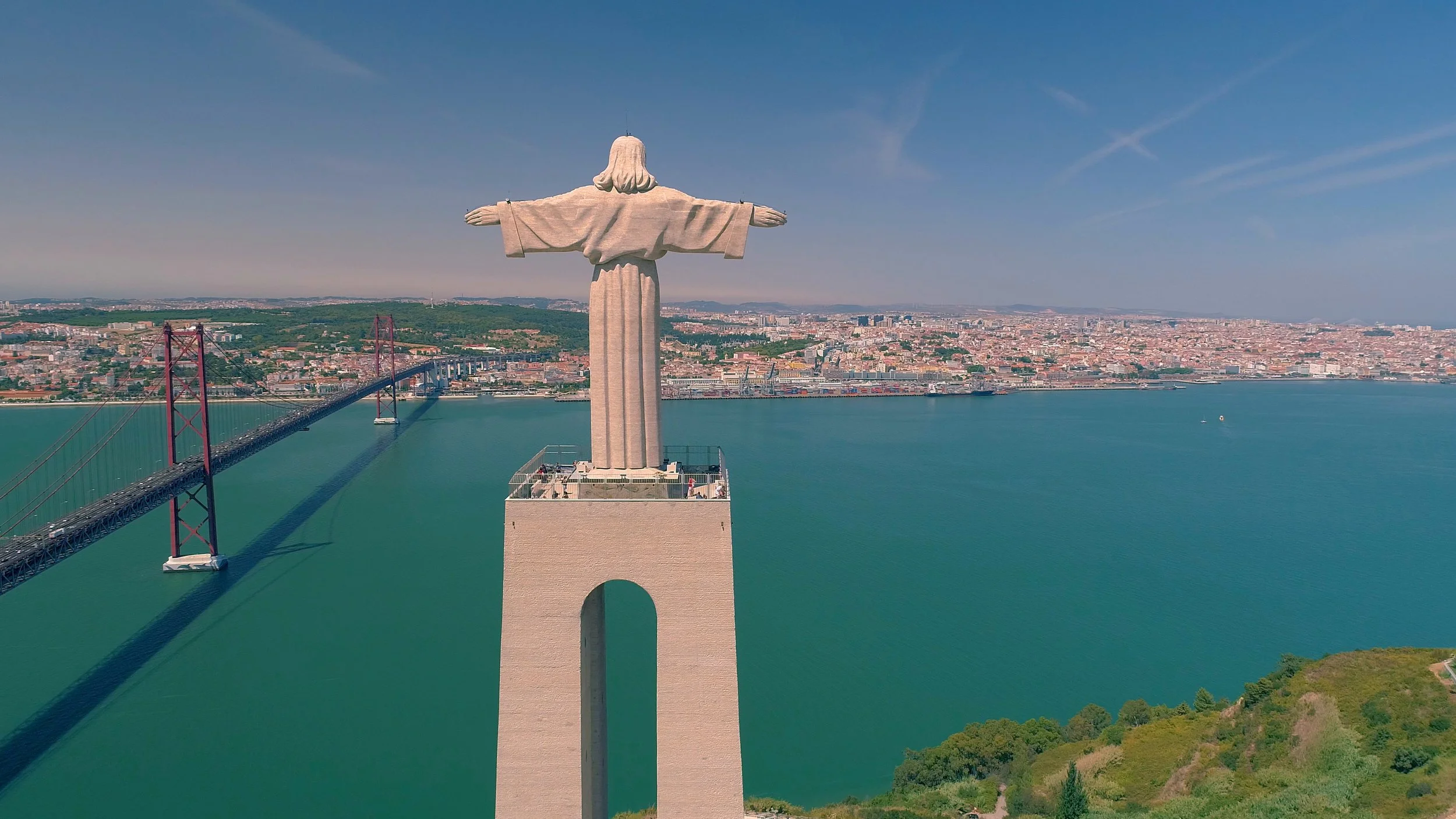 A film and photo location in Portugal from walnut films, An aerial view of the Christ the King statue overlooking Lisbon, Portugal, with the 25 de Abril Bridge crossing the river underneath and the cityscape in the background.