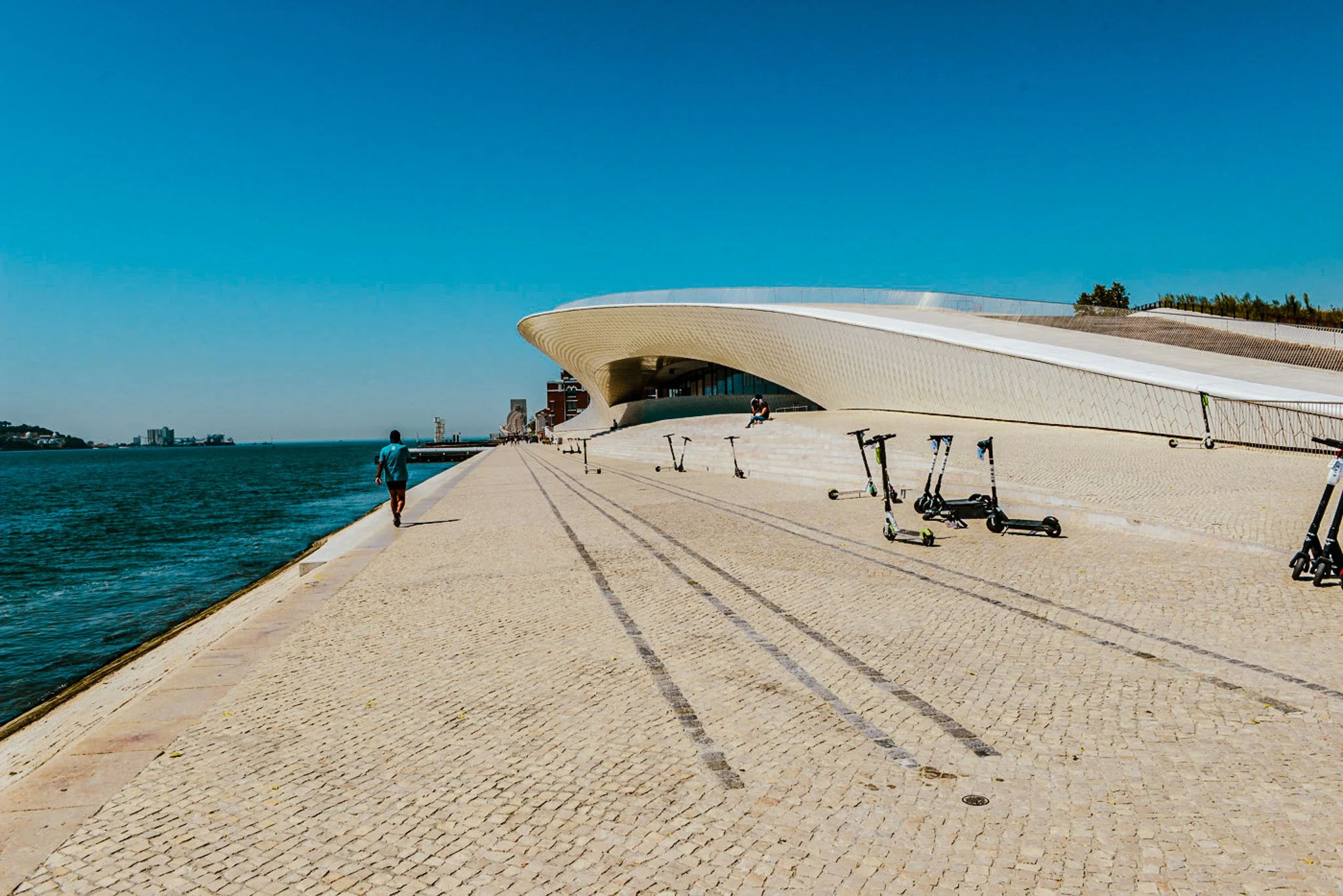 A film and photo location in Portugal from walnut films, A modern building with a curved, futuristic design by a waterfront, with some people walking and electric scooters parked along the cobblestone promenade on a sunny day.