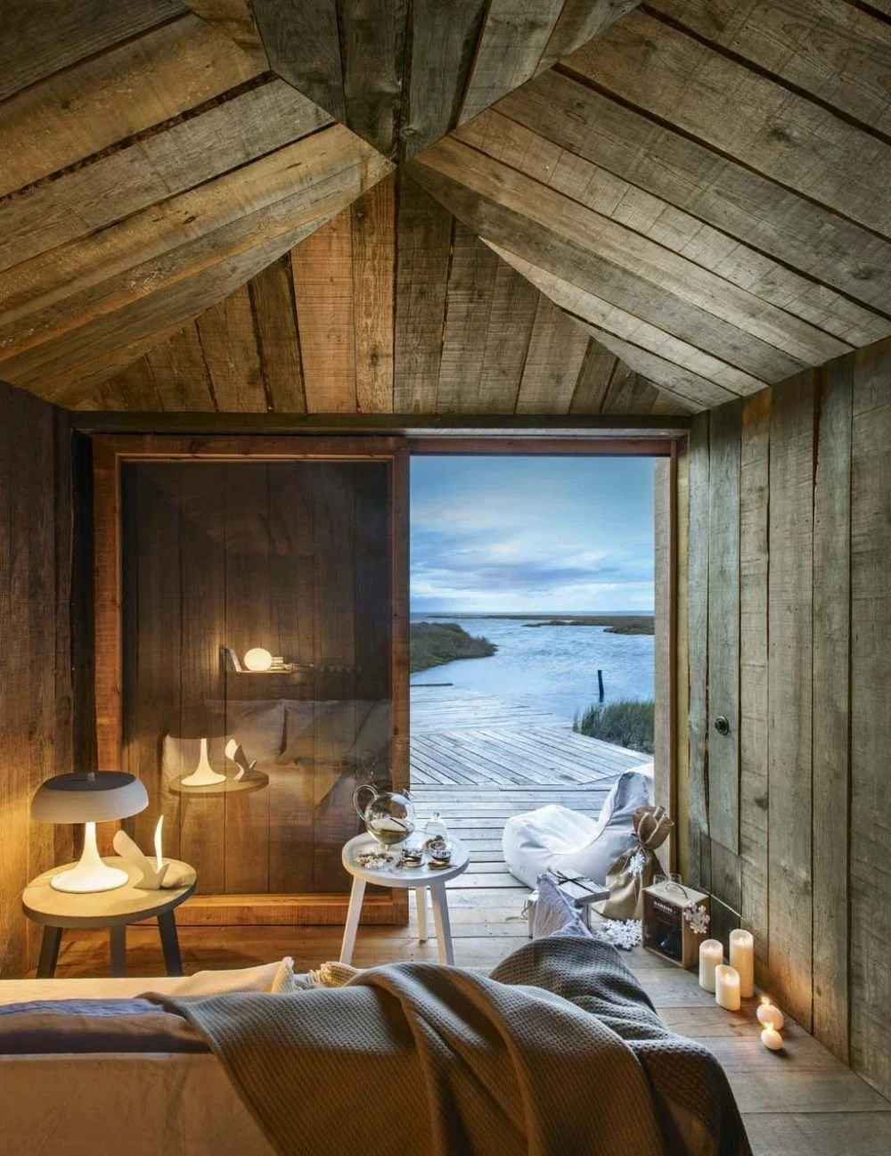 A film and photo location in Portugal from walnut films, Cozy bedroom with wood-paneled walls and ceiling, a glimpse of a deck and water outside through an open door, bedside table with a lamp, candles on the floor, and a beanbag chair.