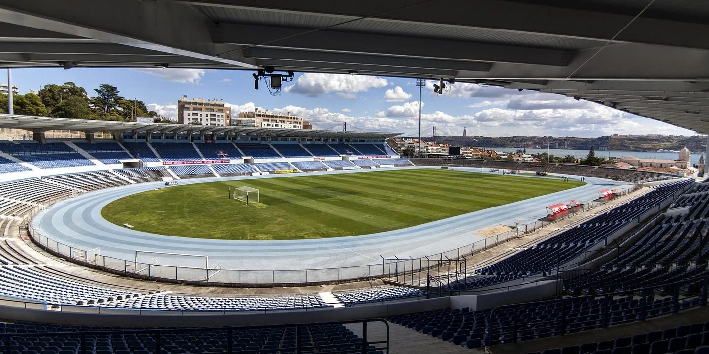 A film and photo location in Portugal from walnut films, Empty outdoor stadium with a running track, green grass field, and blue seats. Overhead roof, buildings, water, and sky in the background.