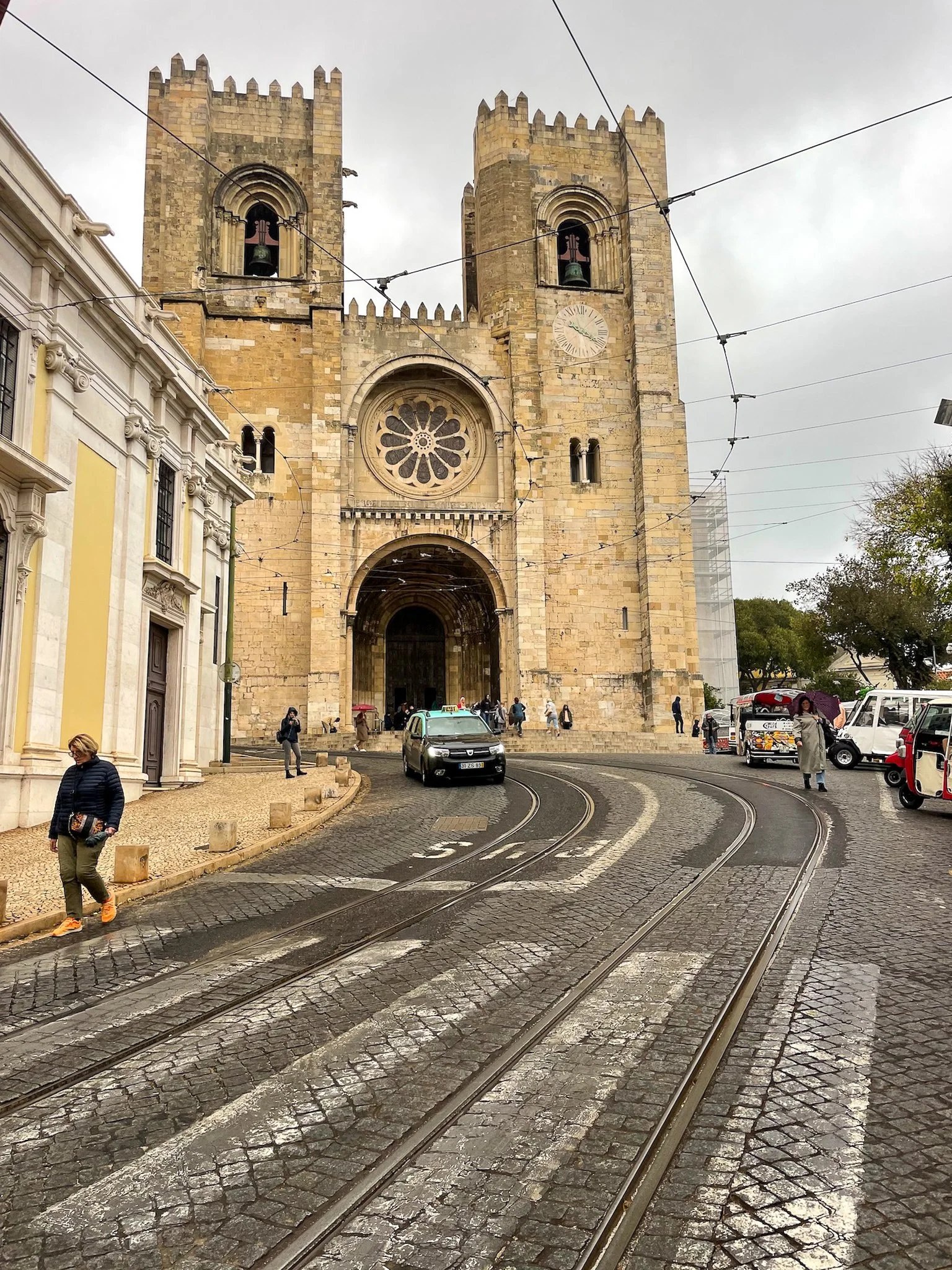A film and photo location in Portugal from walnut films, A historic stone church with twin towers, large round stained glass window, and a clock on the right tower. There are cobblestone streets with tram tracks in front, cars, 