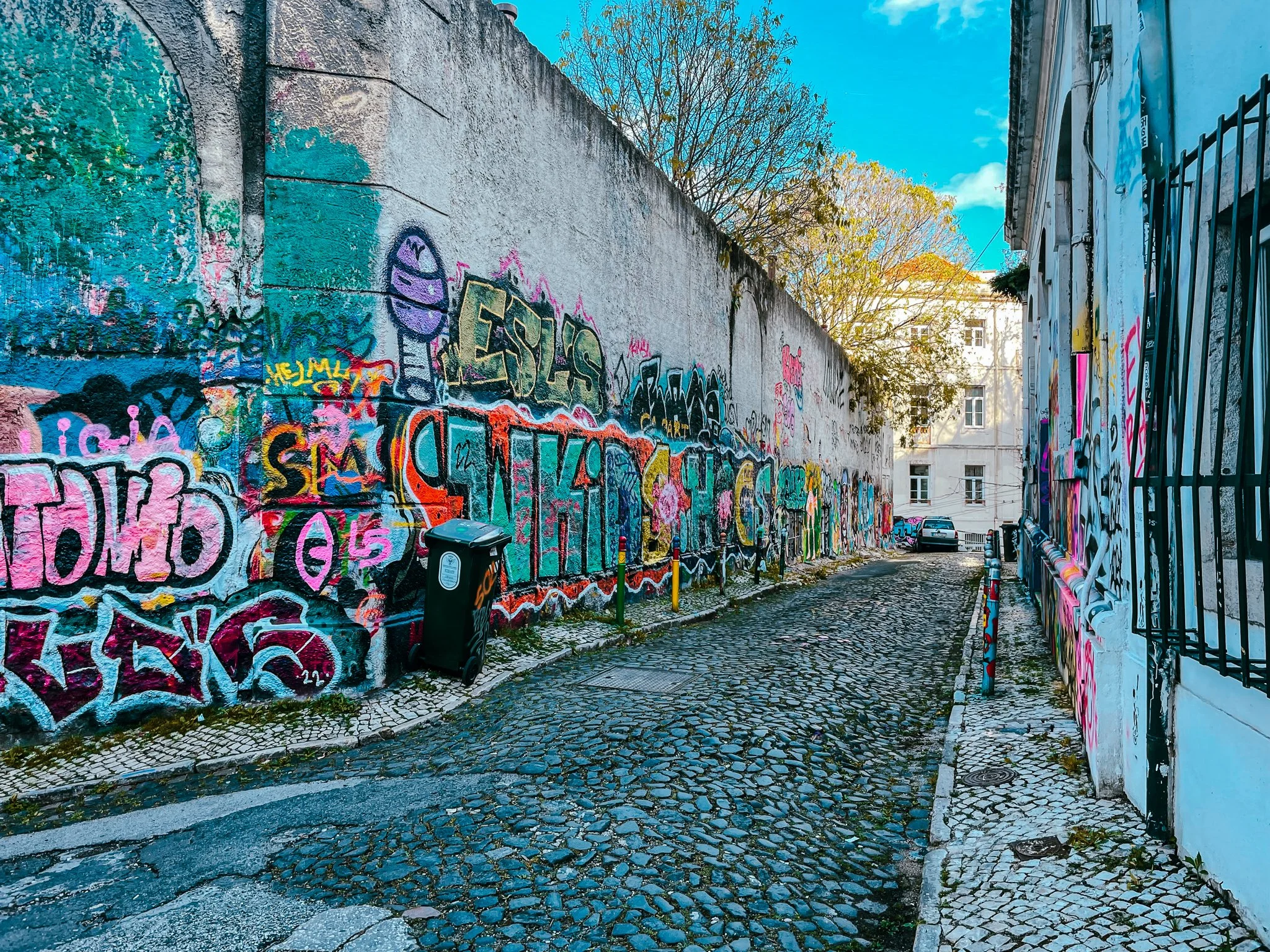 A film and photo location in Portugal from walnut films, A narrow cobblestone street lined with a graffiti-covered wall on the left and a building with a barred window on the right. A black trash bin is in front of the wall