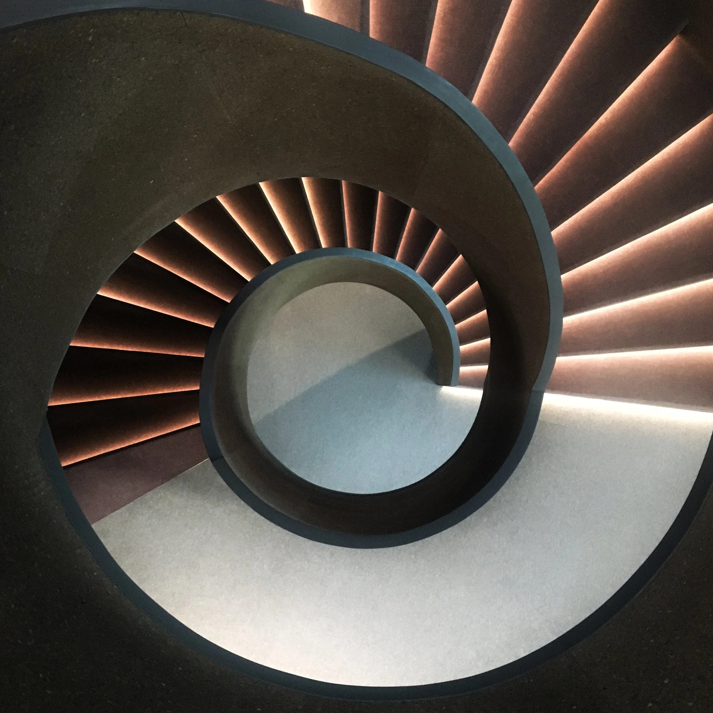 A film and photo location in Portugal from walnut films, A view looking down a spiral staircase with dark steps and a curved handrail, illuminated by warm lighting along the edges of each step.
