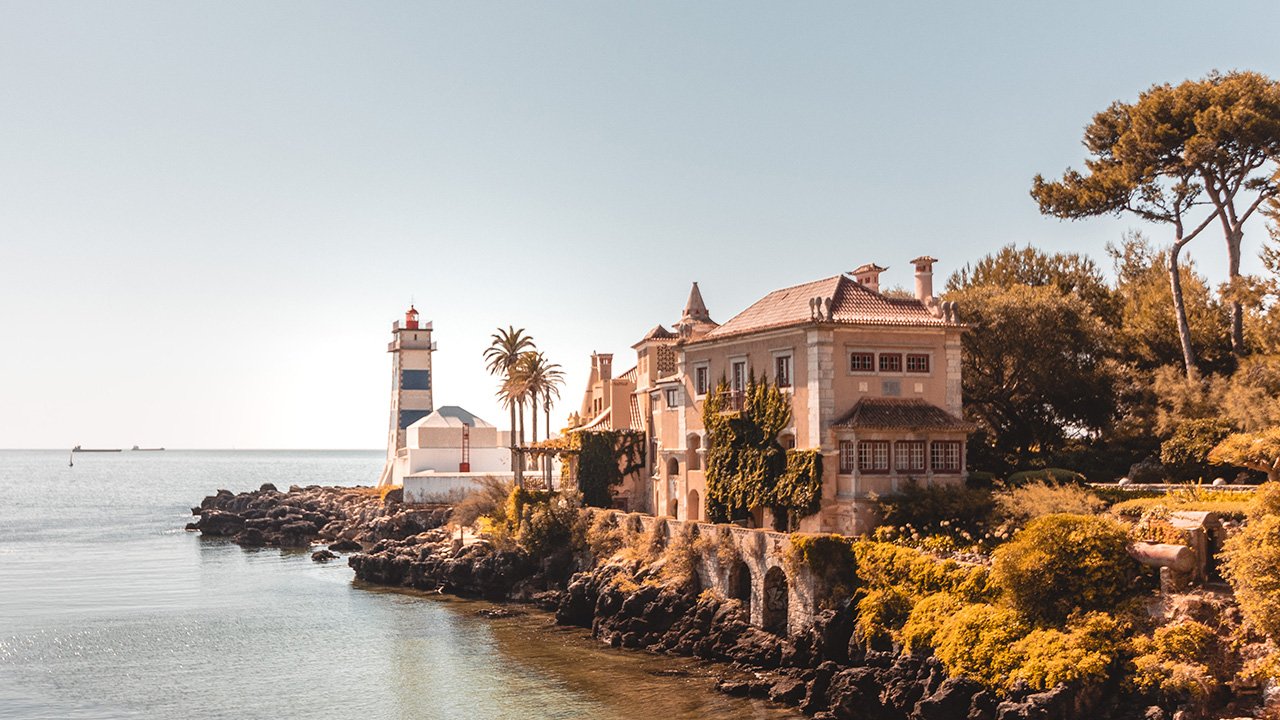 A film and photo location in Portugal from walnut films, Coastal scene with a lighthouse, a historic house, trees, and the ocean under a clear sky.