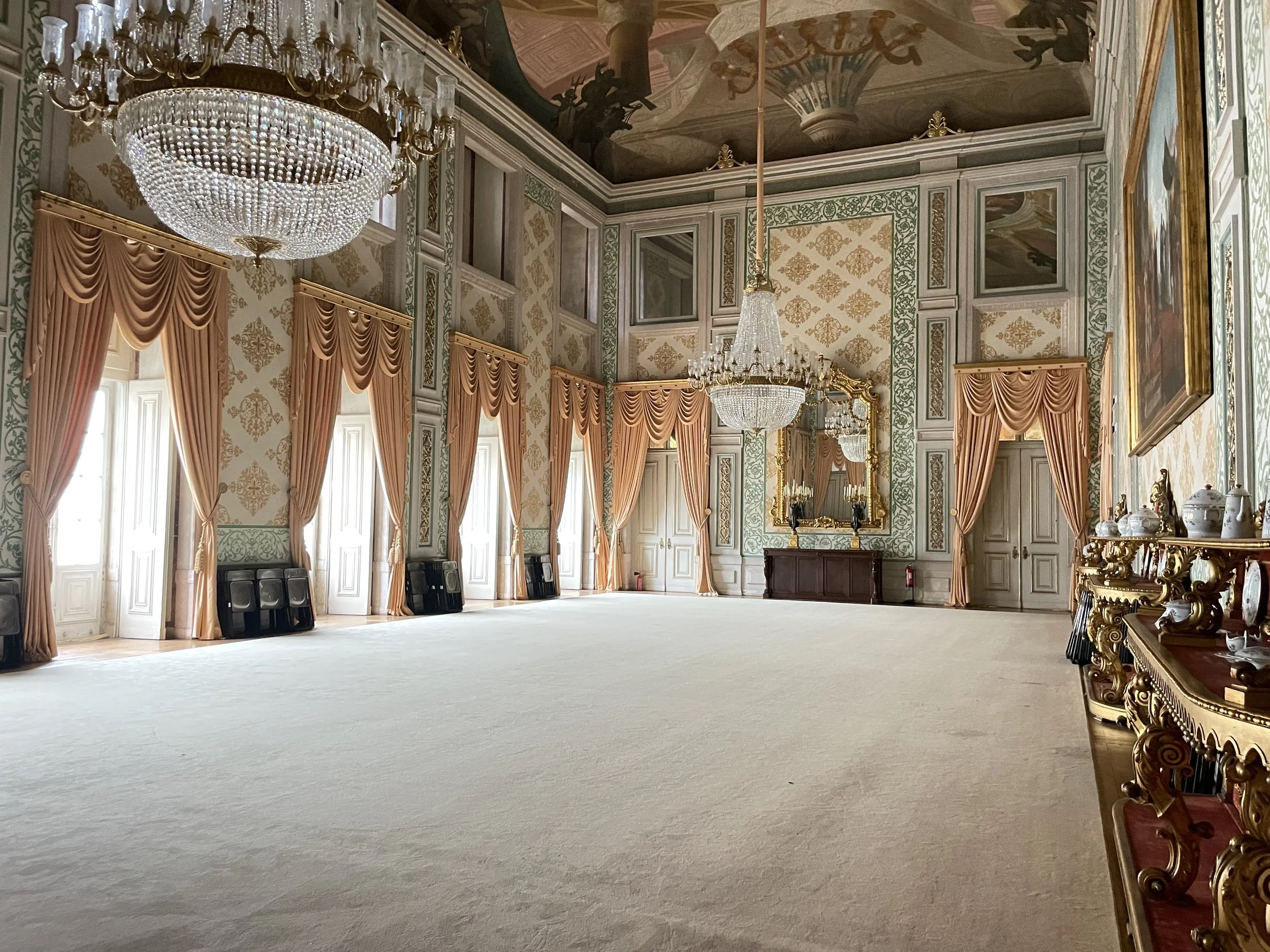 A film and photo location in Portugal from walnut films, An elegant, ornate room with tall windows draped with peach-colored curtains, gilded mirrors, elaborate ceiling paintings, and multiple chandeliers hanging from a richly decorated ceiling.