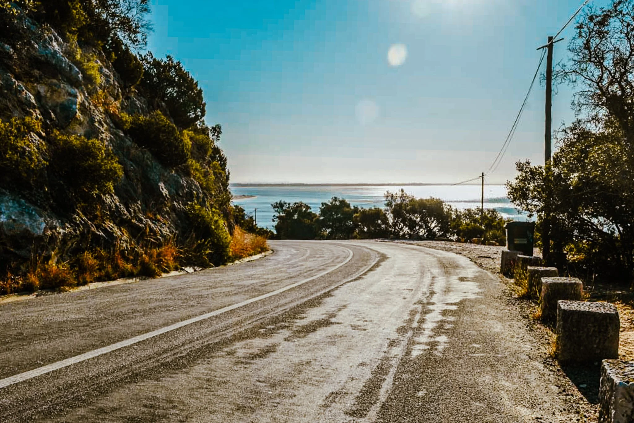 A film and photo location in Portugal from walnut films, A winding dirt road runs along a hillside on the left with trees and shrubs, and next to power lines on the right, leading toward a body of water in the distance under a clear blue sky.