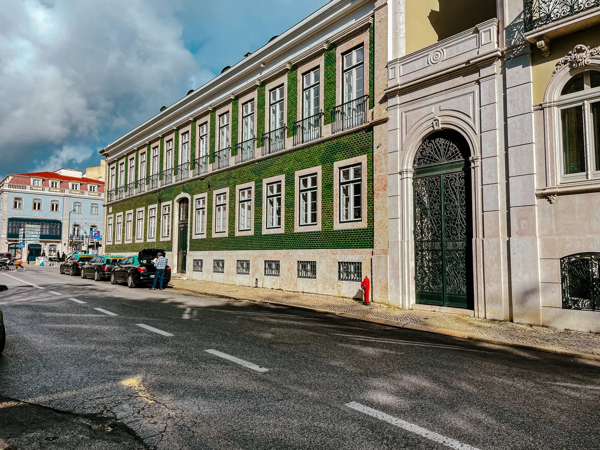 A film and photo location in Portugal from walnut films, Street scene with parked cars, people walking, and a historic building with green tiles and ornate entrance in an urban area.