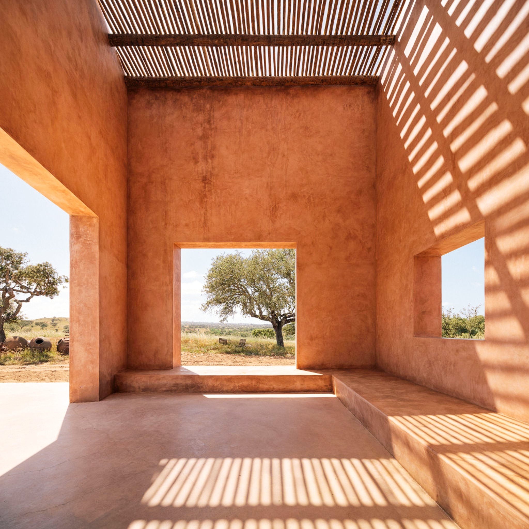 A film and photo location in Portugal from walnut films, View from inside a structure with reddish walls and wooden beams, showing shadows of slats on the ceiling and floor, with a landscape of trees and open sky visible through large open windows.