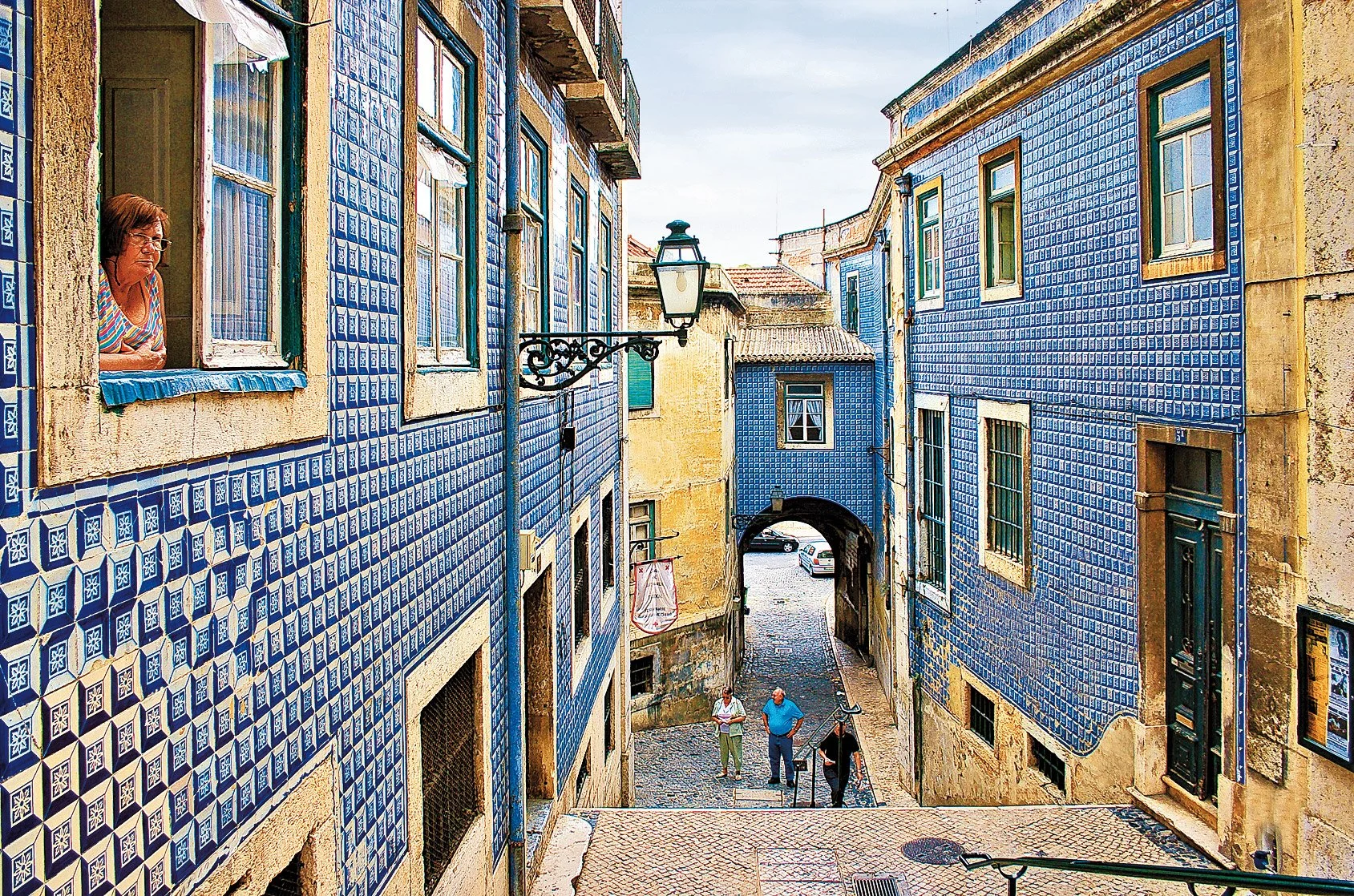 A film and photo location in Portugal from walnut films, Street view of buildings with blue and yellow tiled facades, arched passageway at the end, people walking and looking out the window of the building on the left.