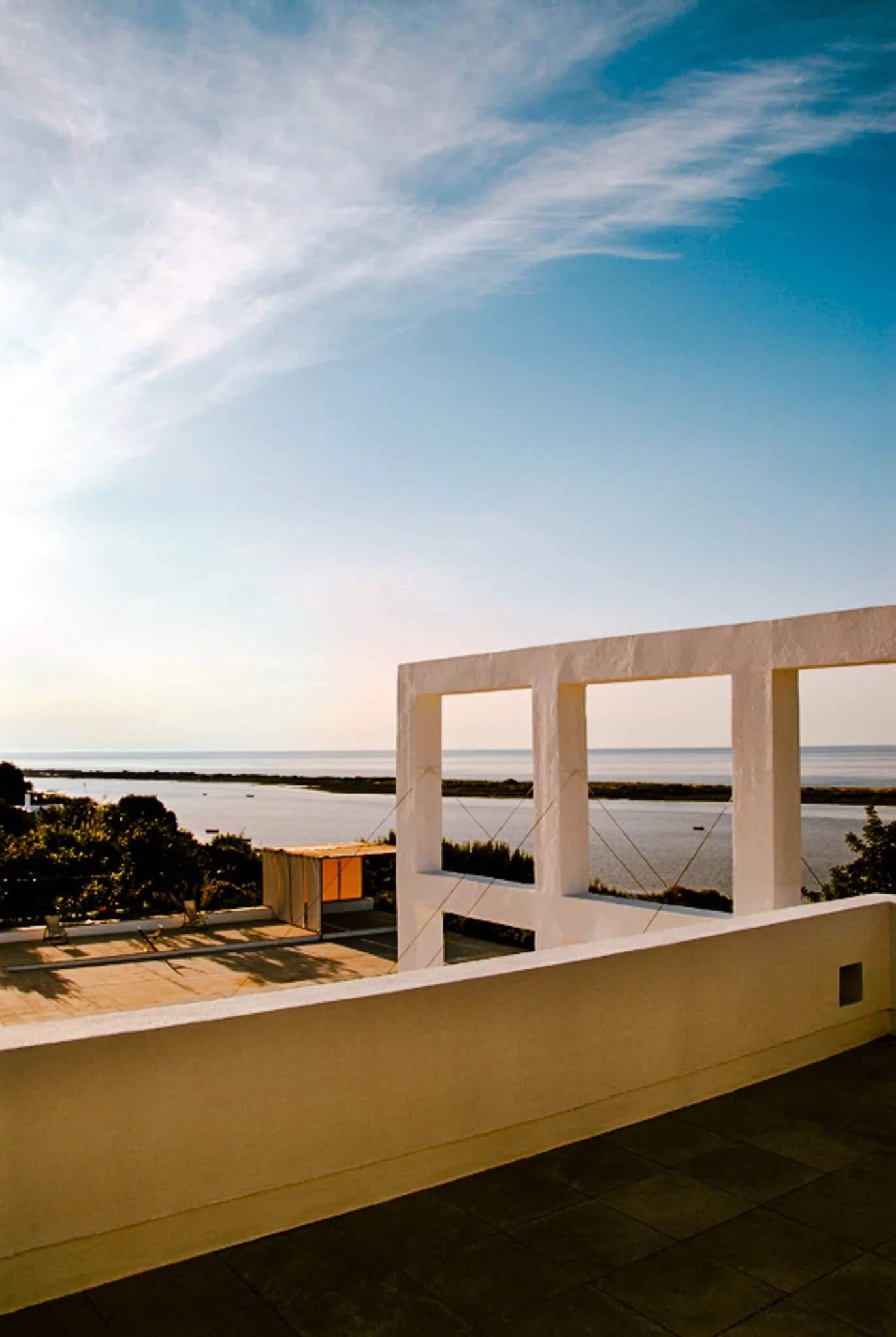 A film and photo location in Portugal from walnut films, View of a seaside landscape during sunset, with a white architectural structure in the foreground, trees, water, and a partly cloudy sky.