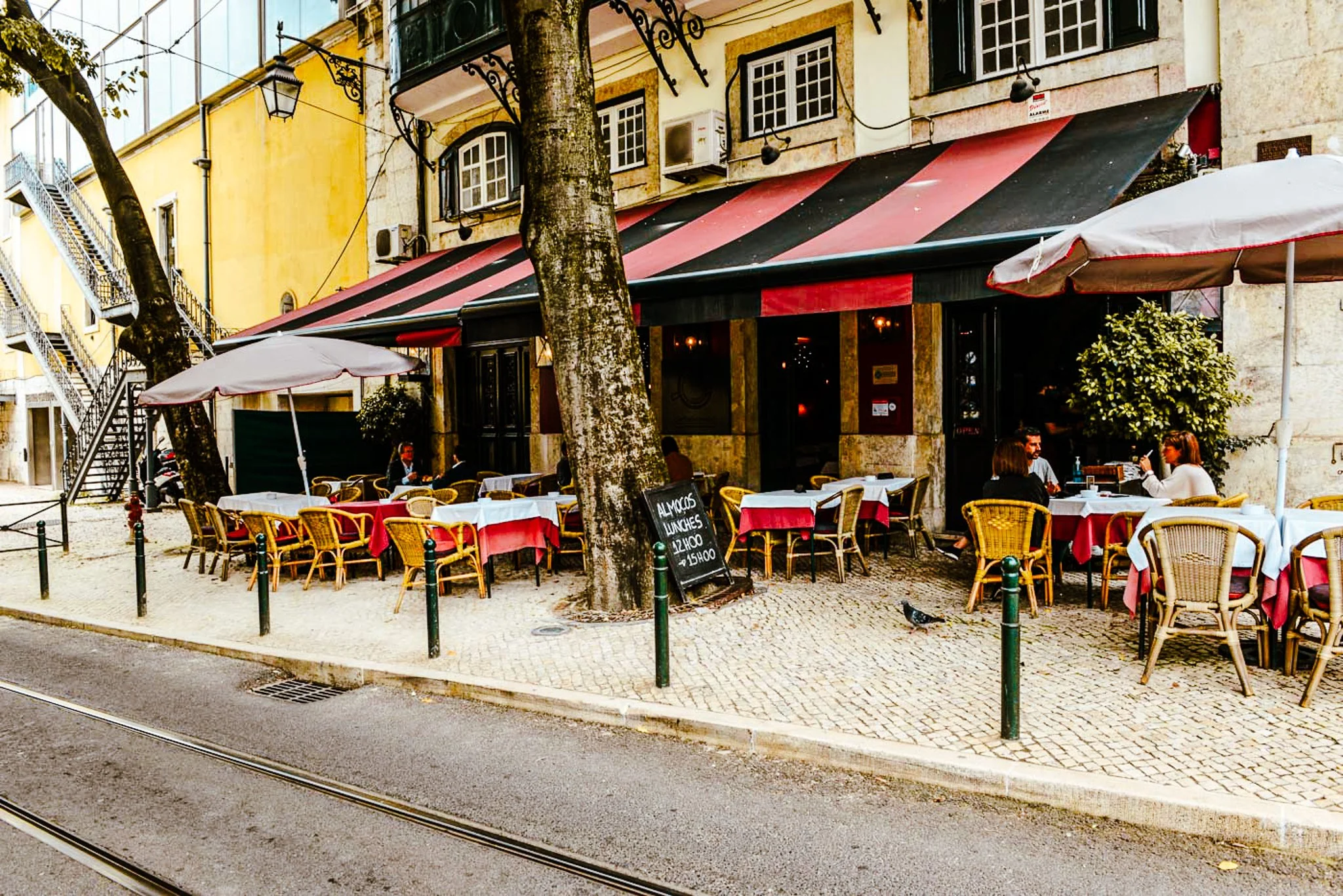 A film and photo location in Portugal from walnut films, Outdoor patio of a restaurant with red and black striped awning, tables with red tablecloths, umbrellas, and people sitting and dining.