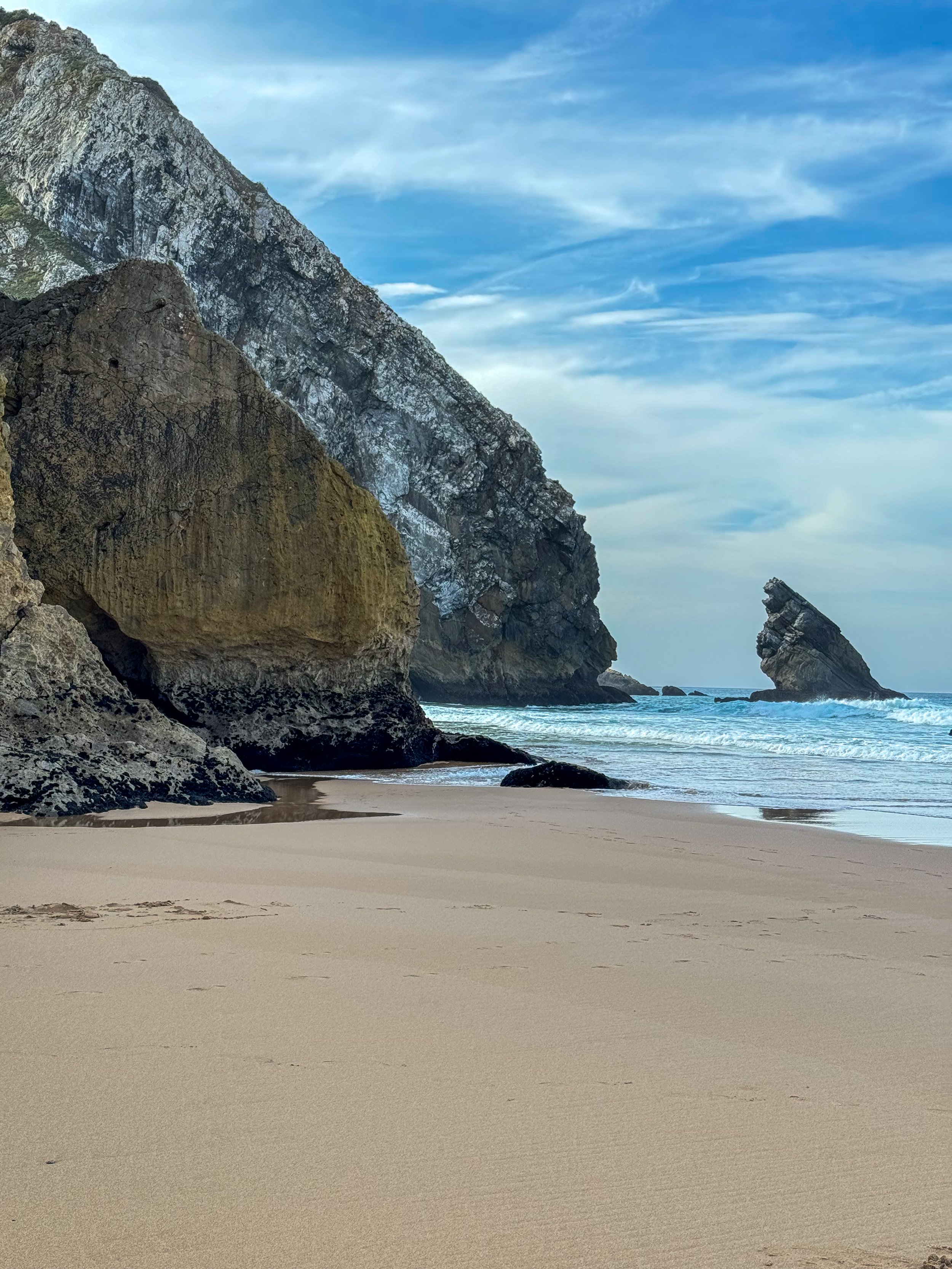 A film and photo location in Portugal, A sandy beach with large rocks and cliffs in the background, under a cloudy sky.