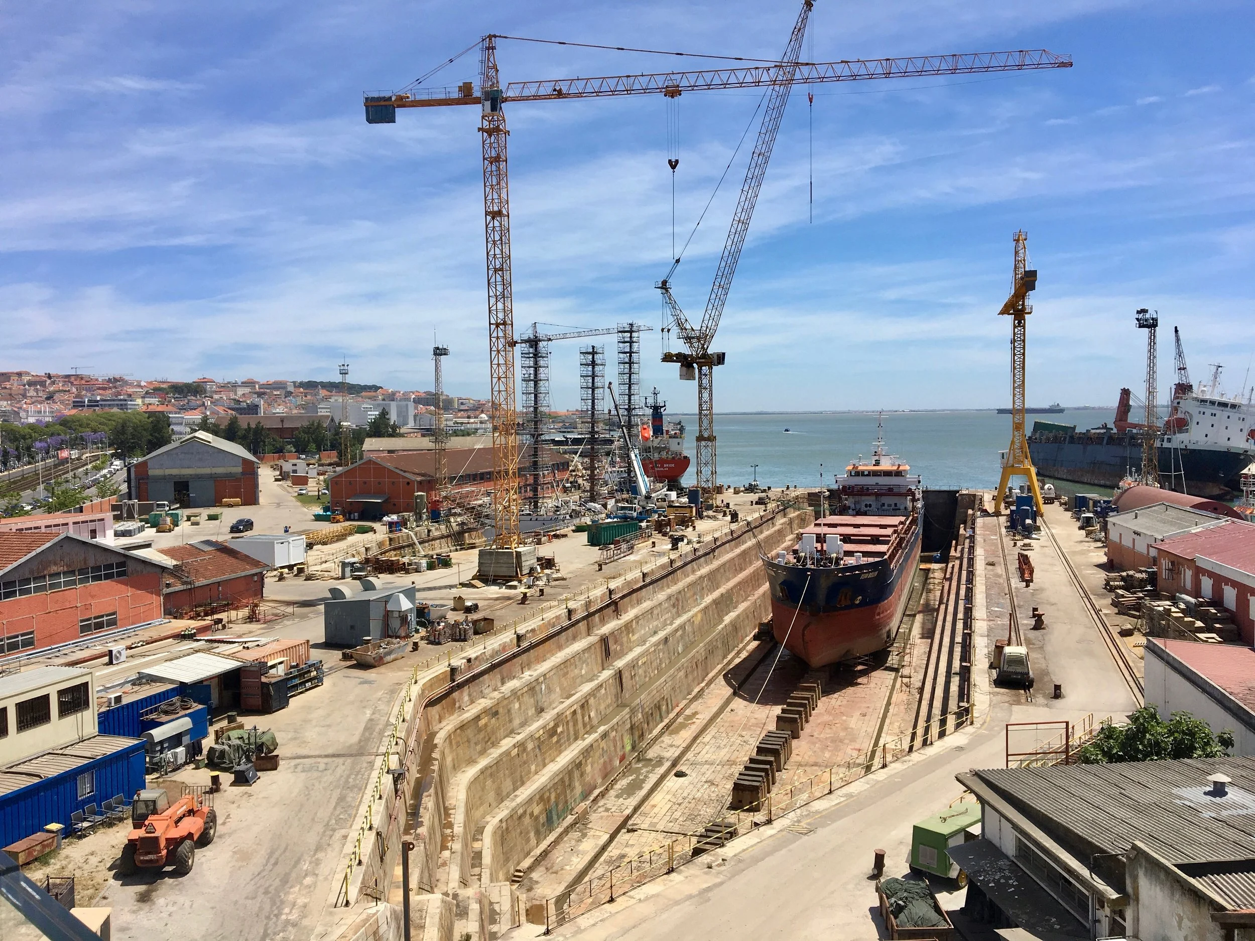 A film and photo location in Portugal from walnut films, View of a busy shipyard with cranes, boats, and buildings by the water, under a partly cloudy sky.