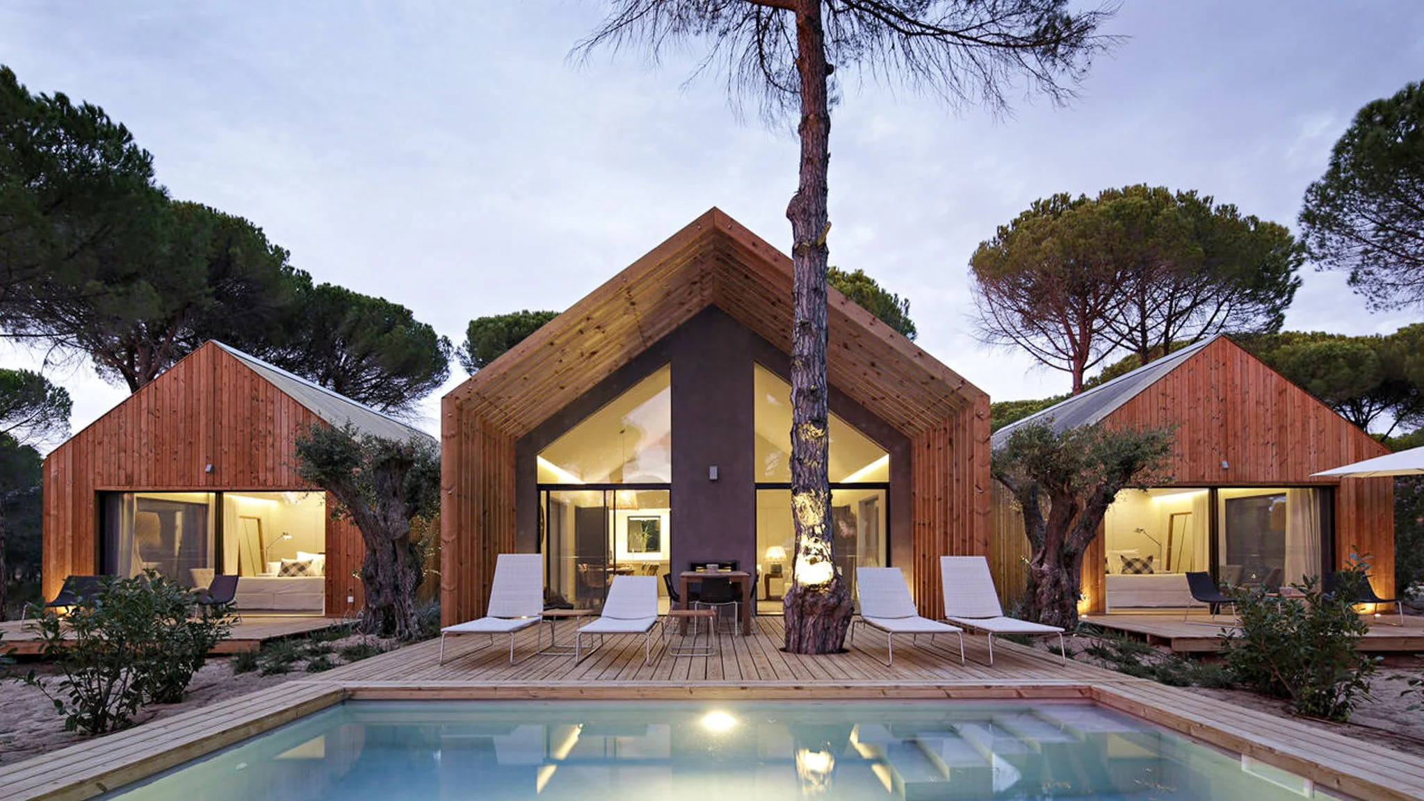 A film and photo location in Portugal from walnut films, Modern wooden house with large glass windows, outdoor deck with lounge chairs, surrounded by trees, with a small pool in front, and interior lighting visible through the windows.