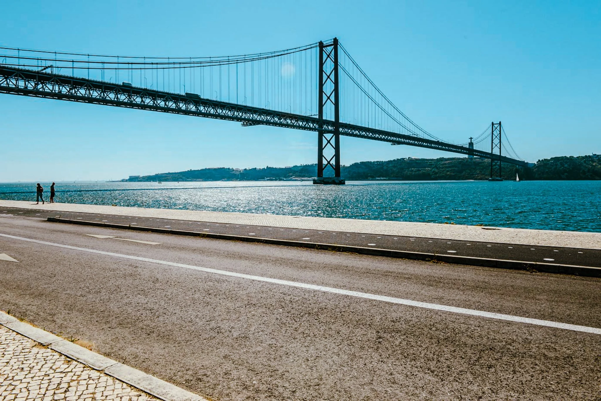 A film and photo location in Portugal from walnut films, View of a large suspension bridge over a body of water, with two people walking along a paved promenade in the foreground and a sailboat in the distance.