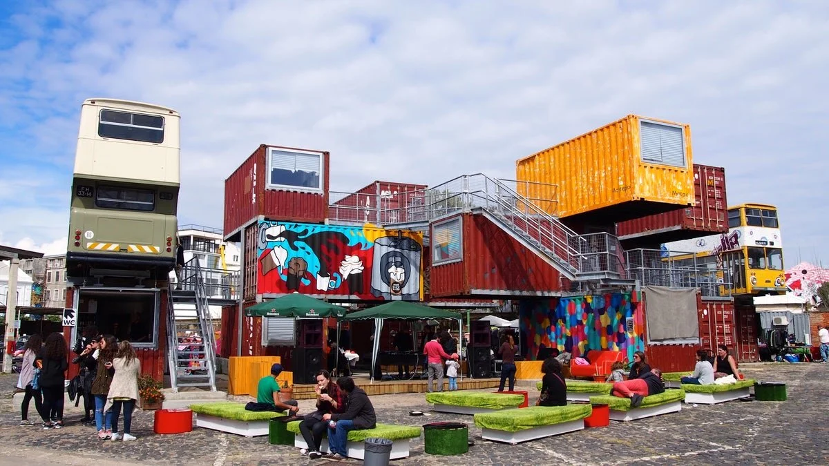 A film and photo location in Portugal from walnut films, People sitting and talking outside a colorful, industrial-style building made of stacked shipping containers, with murals and windows, in an open public space.