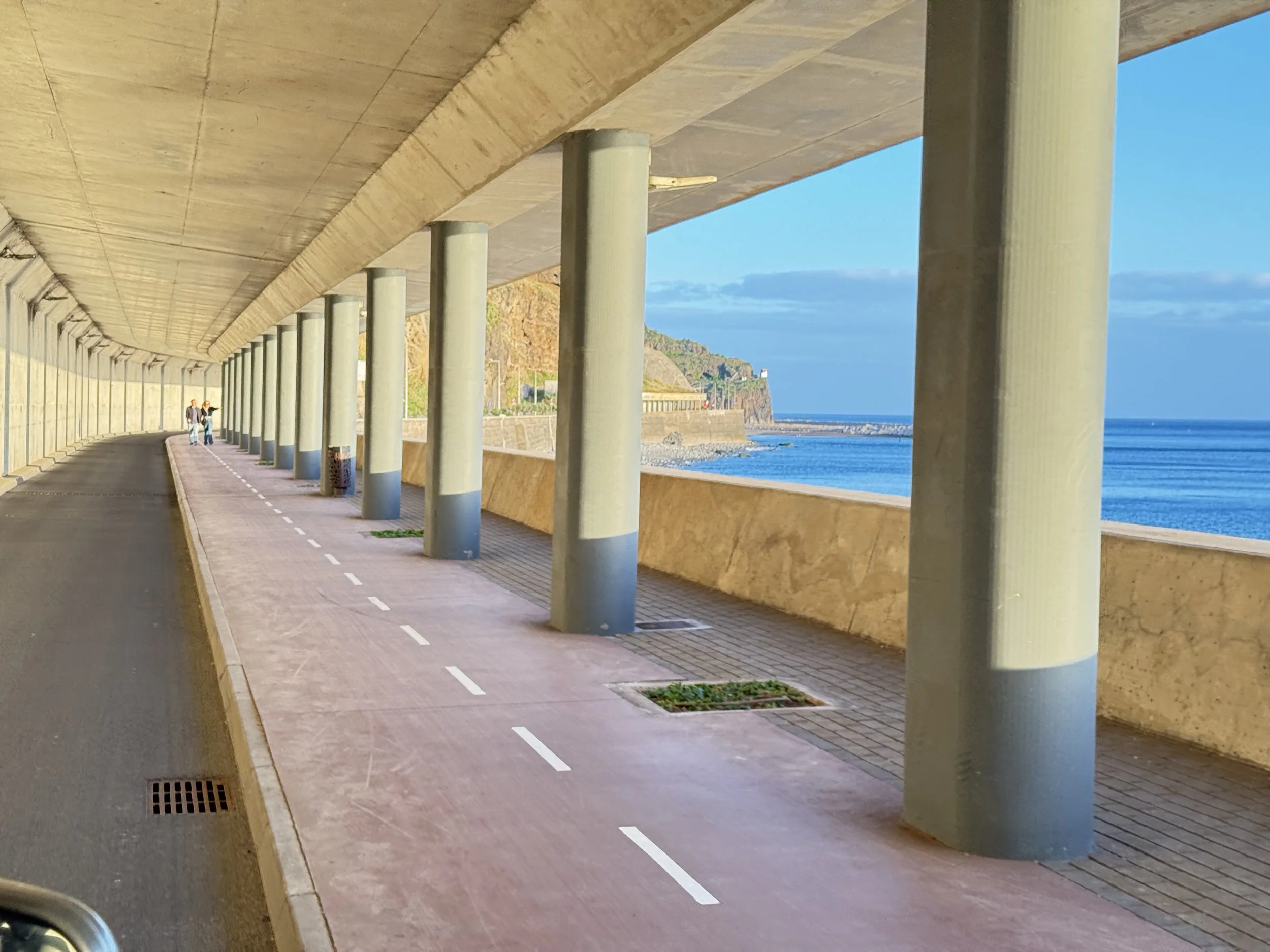 A film and photo location in Portugal from walnut films, Underpass with a curved ceiling and large green support pillars, a pathway with a dashed line, and a view of a coastline with hills and a lighthouse in the distance.