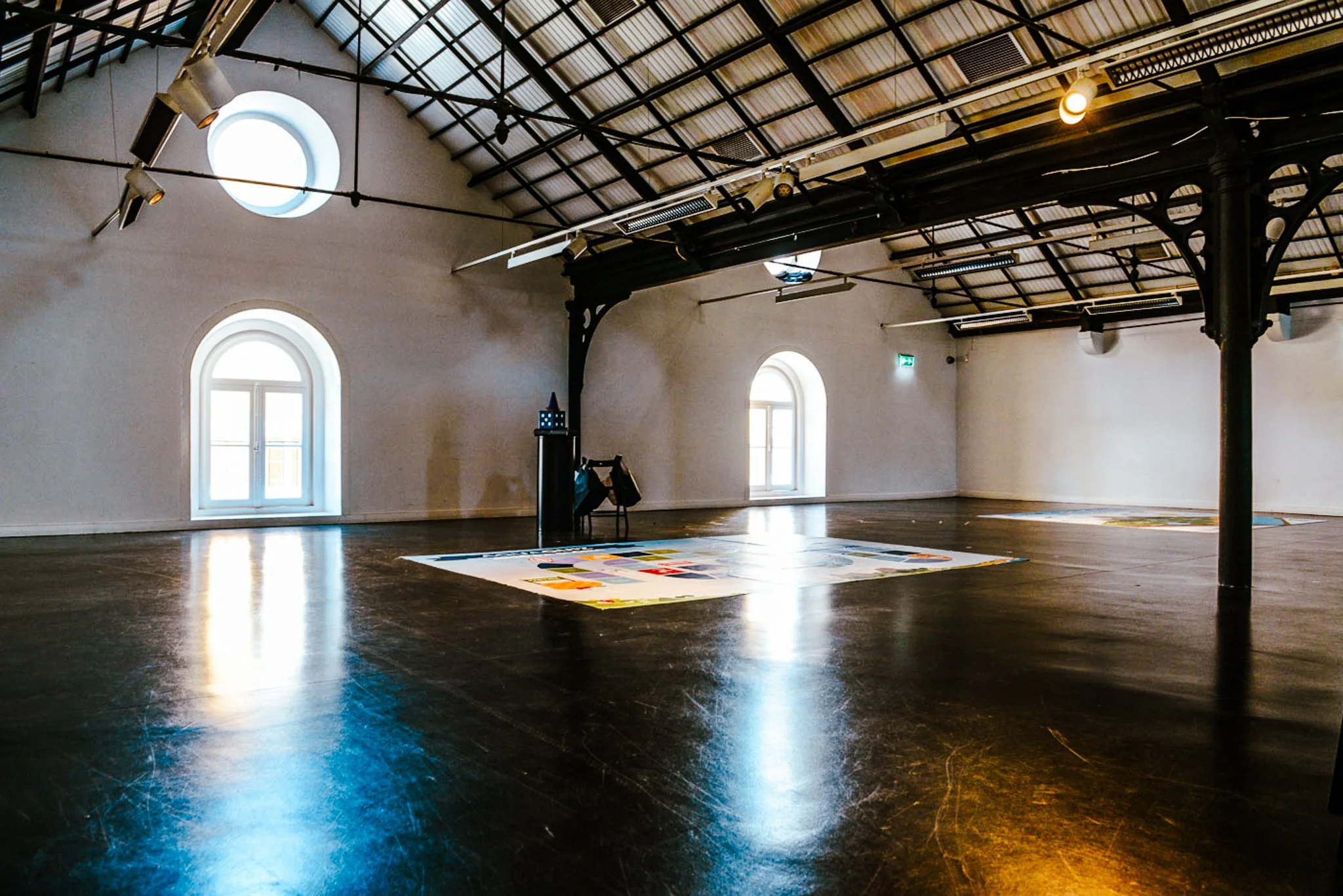 A film and photo location in Portugal from walnut films, Empty art gallery with arched windows, wooden floors, and black steel beams supporting a metal roof. There is a colorful artwork on the floor and two small art pieces on a stand in the corner.
