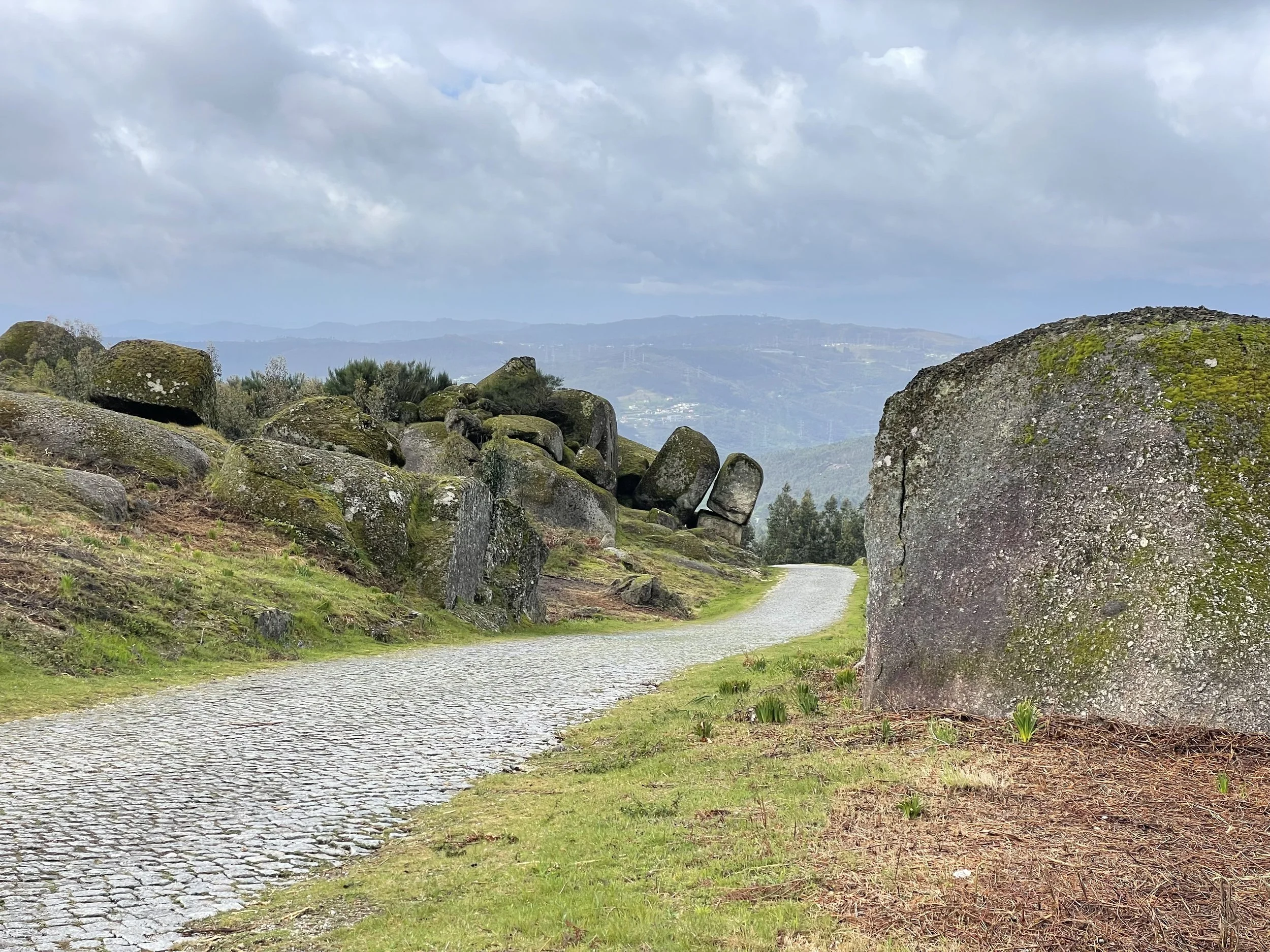 A film and photo location in Portugal from walnut films, A gravel pathway winding through a lush green landscape with moss-covered rocks on each side. In the background, there are rolling hills under a cloudy sky.