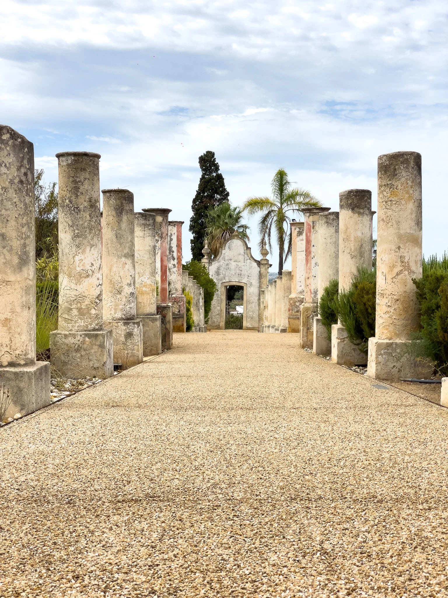 A film and photo location in Portugal from walnut films, Pathway bordered by old stone columns leading to a white building with decorative archway, surrounded by palm trees and other vegetation, under a partly cloudy sky.