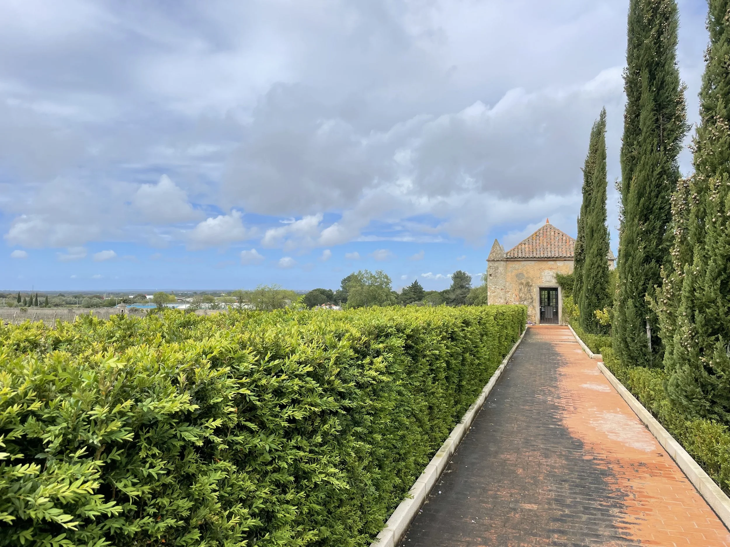 A film and photo location in Portugal from walnut films, A stone building with a tiled roof and a small pointed tower, flanked by tall, narrow cypress trees on a pathway, under a partly cloudy sky.