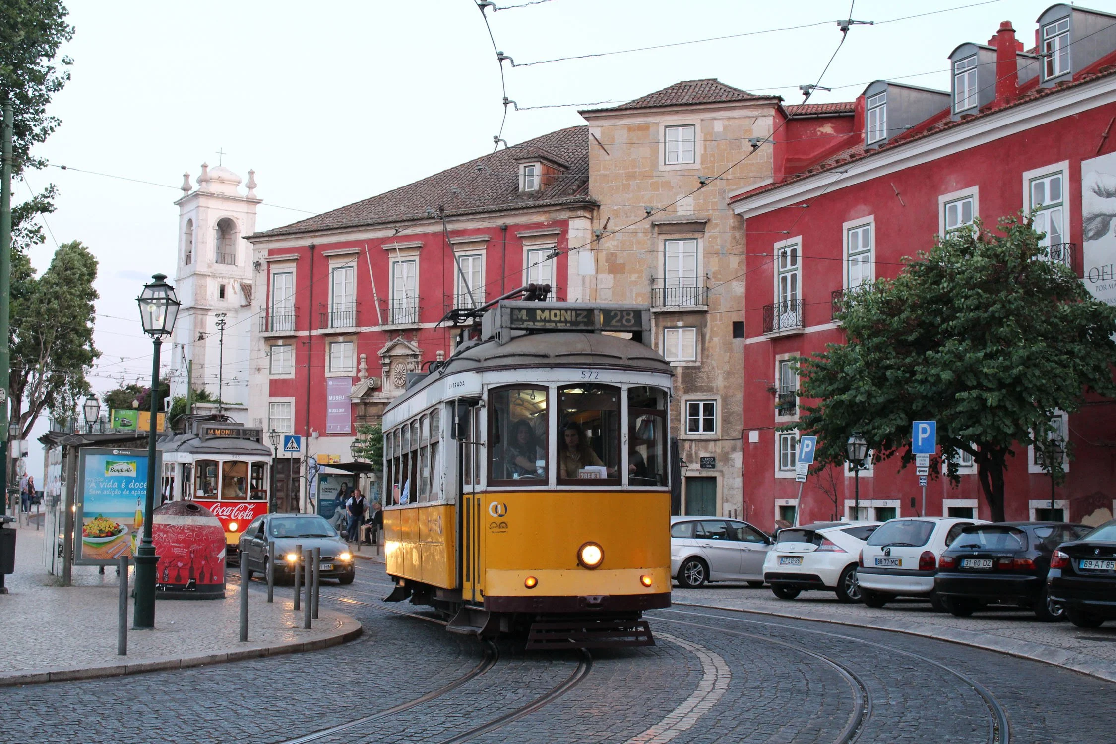 A film and photo location in Portugal from walnut films, Yellow tram moving along curved tracks as other vehicles and pedestrians are present on a city street with historic buildings and overhead electric wires.