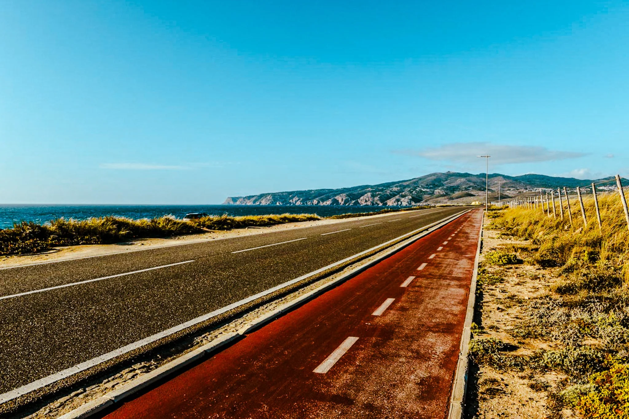 A film and photo location in Portugal from walnut films, Road along the coast with a bike lane, fencing, and hills in the background under a blue sky.