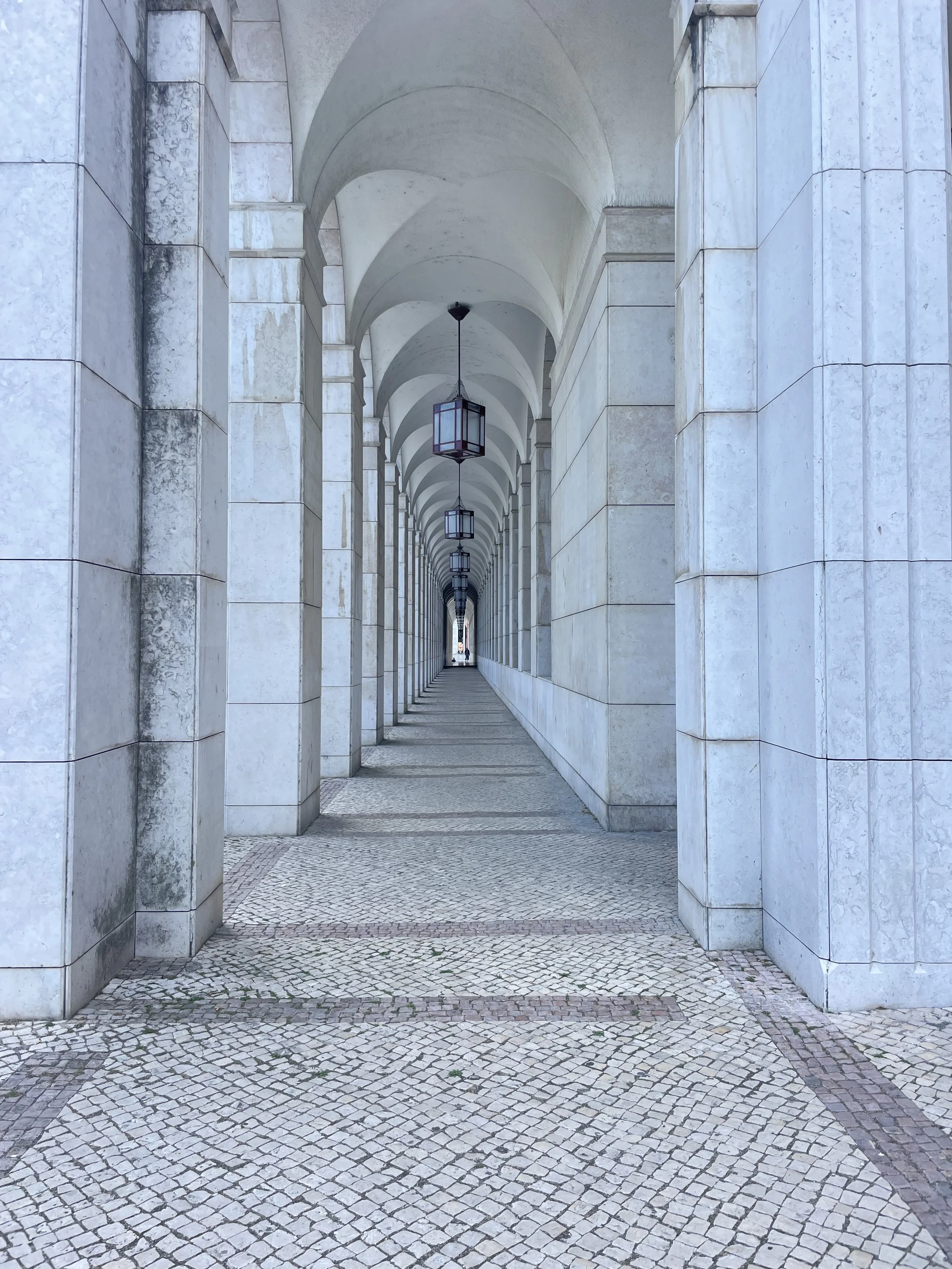 A film and photo location in Portugal from walnut films, A covered walkway with stone arches and columns, lined with hanging lantern lights, and a tiled pavement floor.