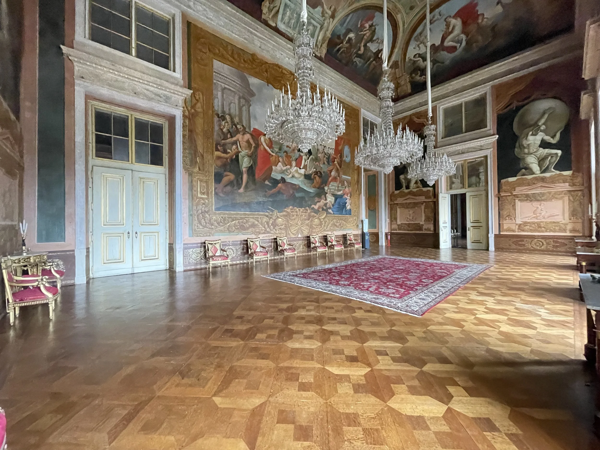 A film and photo location in Portugal from walnut filmAn ornate interior room with large chandeliers hanging from the ceiling, elaborate wall paintings, gold accents, tall windows, a red patterned rug, and a row of red and gold chairs along the wall.