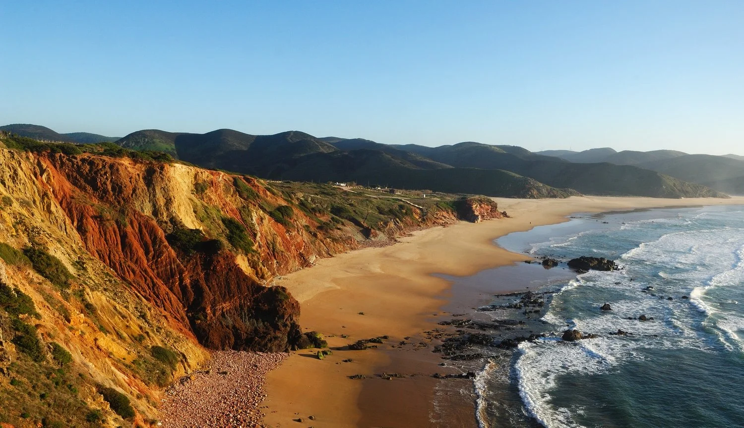 A film and photo location in Portugal, View of a sandy beach with colorful cliffs on the left and mountains in the background, ocean waves crashing onto the shore during sunset.