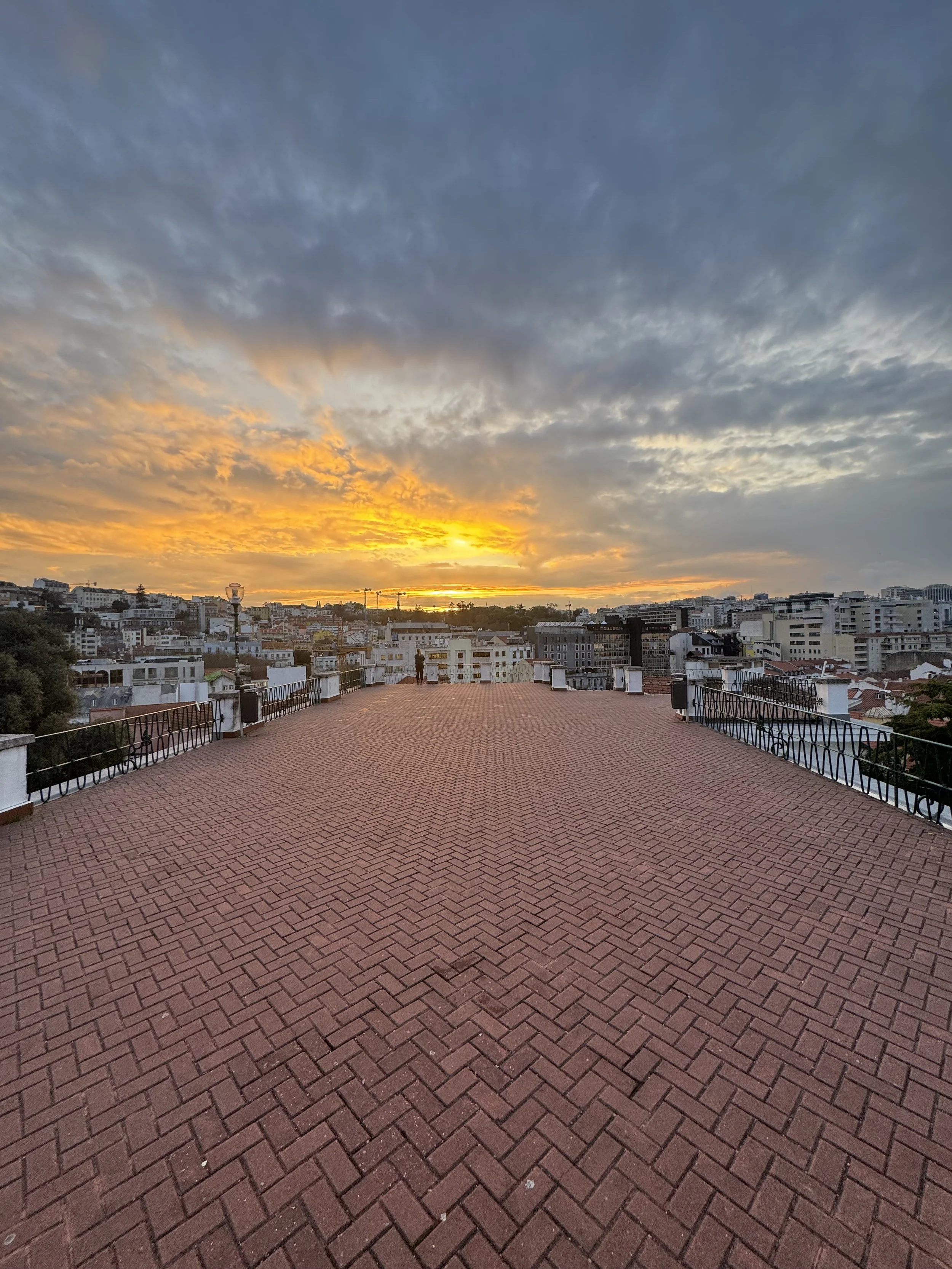 A film and photo location in Portugal from walnut films, A rooftop terrace with a brick floor, black metal railing, and a view of a city skyline at sunset, with a person standing and looking towards the horizon.