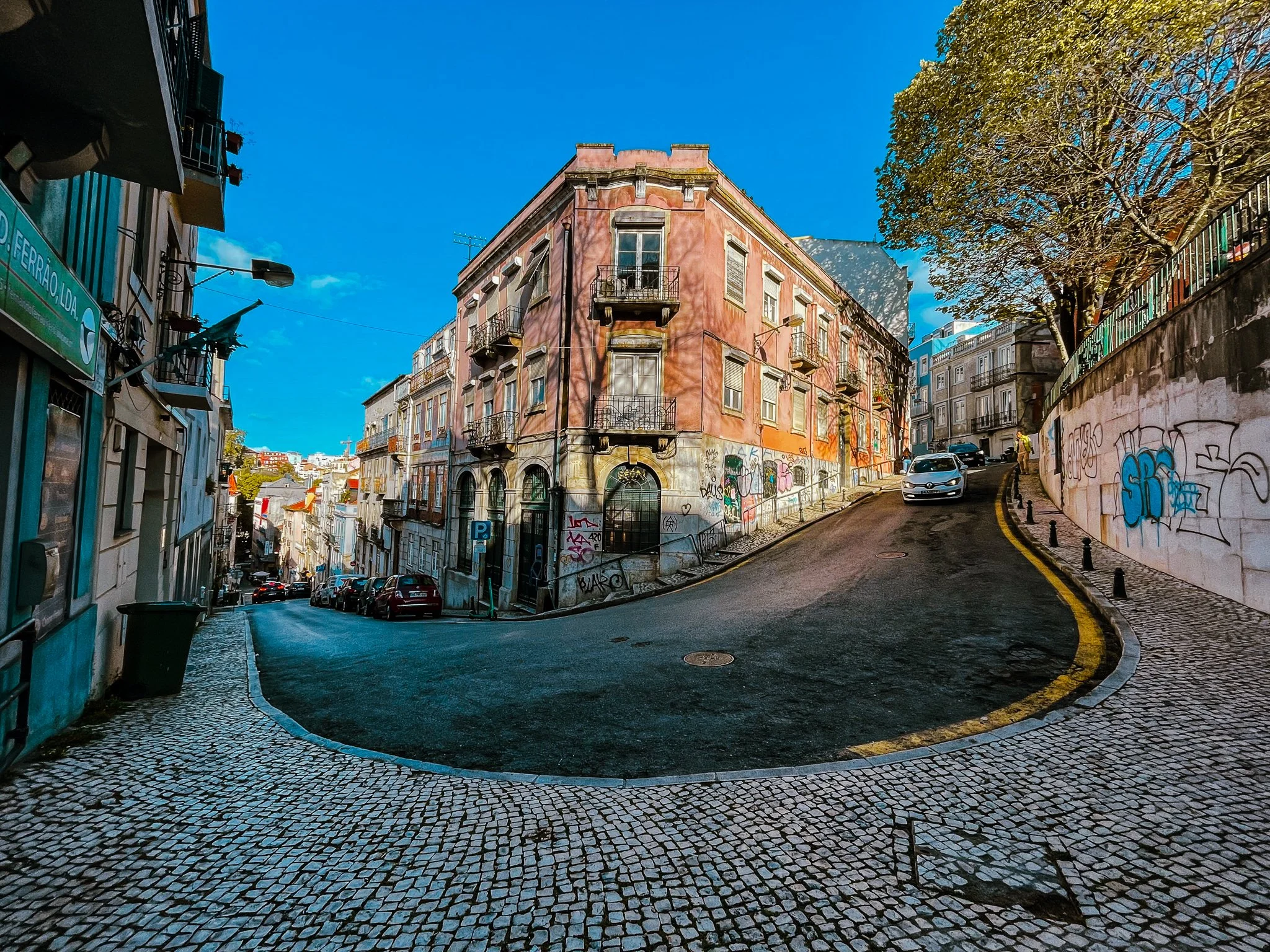 A film and photo location in Portugal from walnut films, A street scene with old buildings, one pink and one white, and a steep hill. The street is paved with cobblestones and has parked cars. 