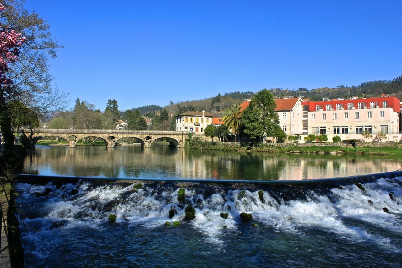 A film and photo location in Portugal from walnut films, A picturesque river scene with a small waterfall in the foreground, a stone bridge with arches crossing the river, and colorful buildings, including a white building with a cross,