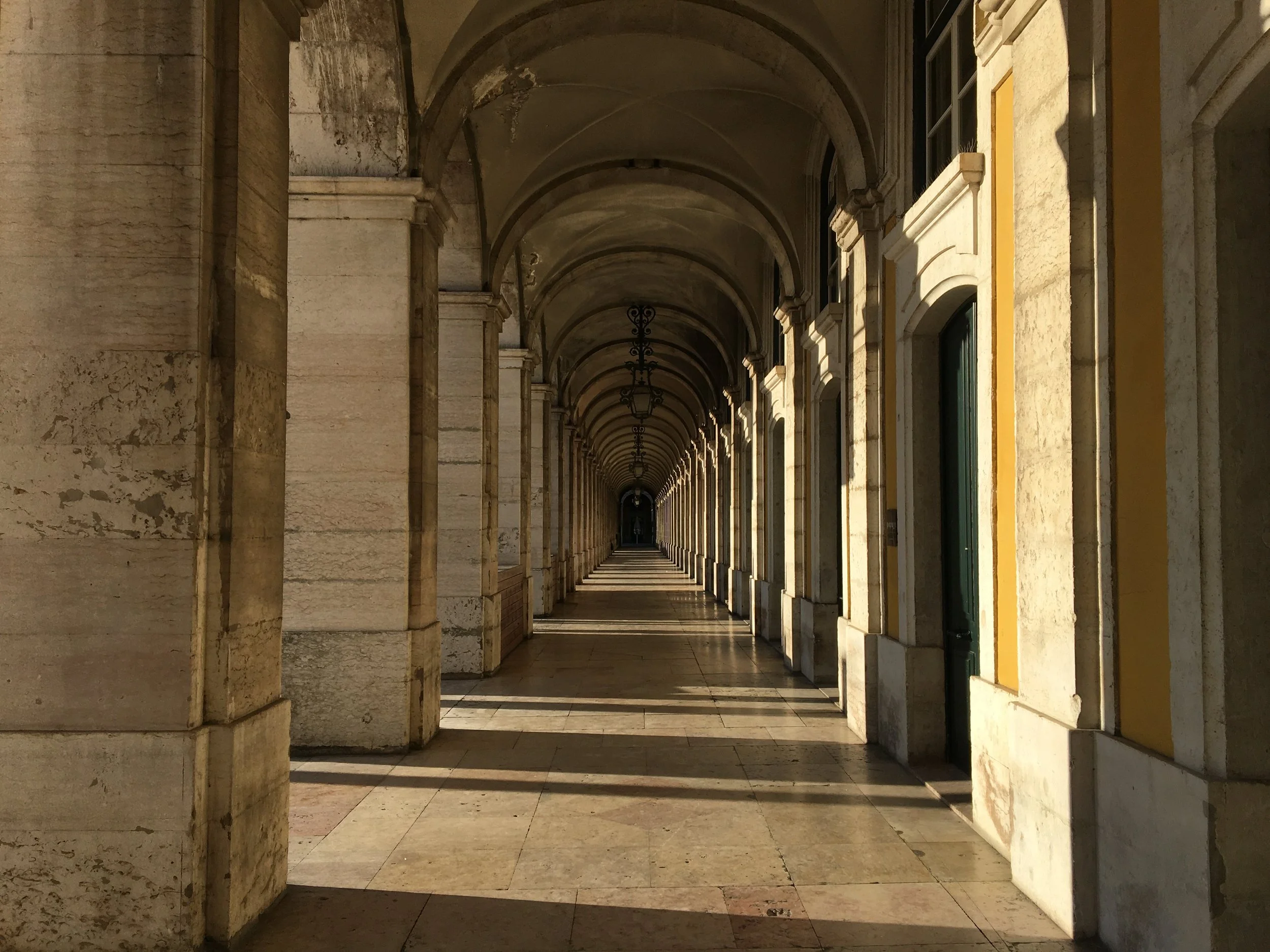 A film and photo location in Portugal from walnut films, A colonnaded walkway with arches, stone pillars, and hanging lanterns, viewed from within a building, with light creating shadows on the floor.