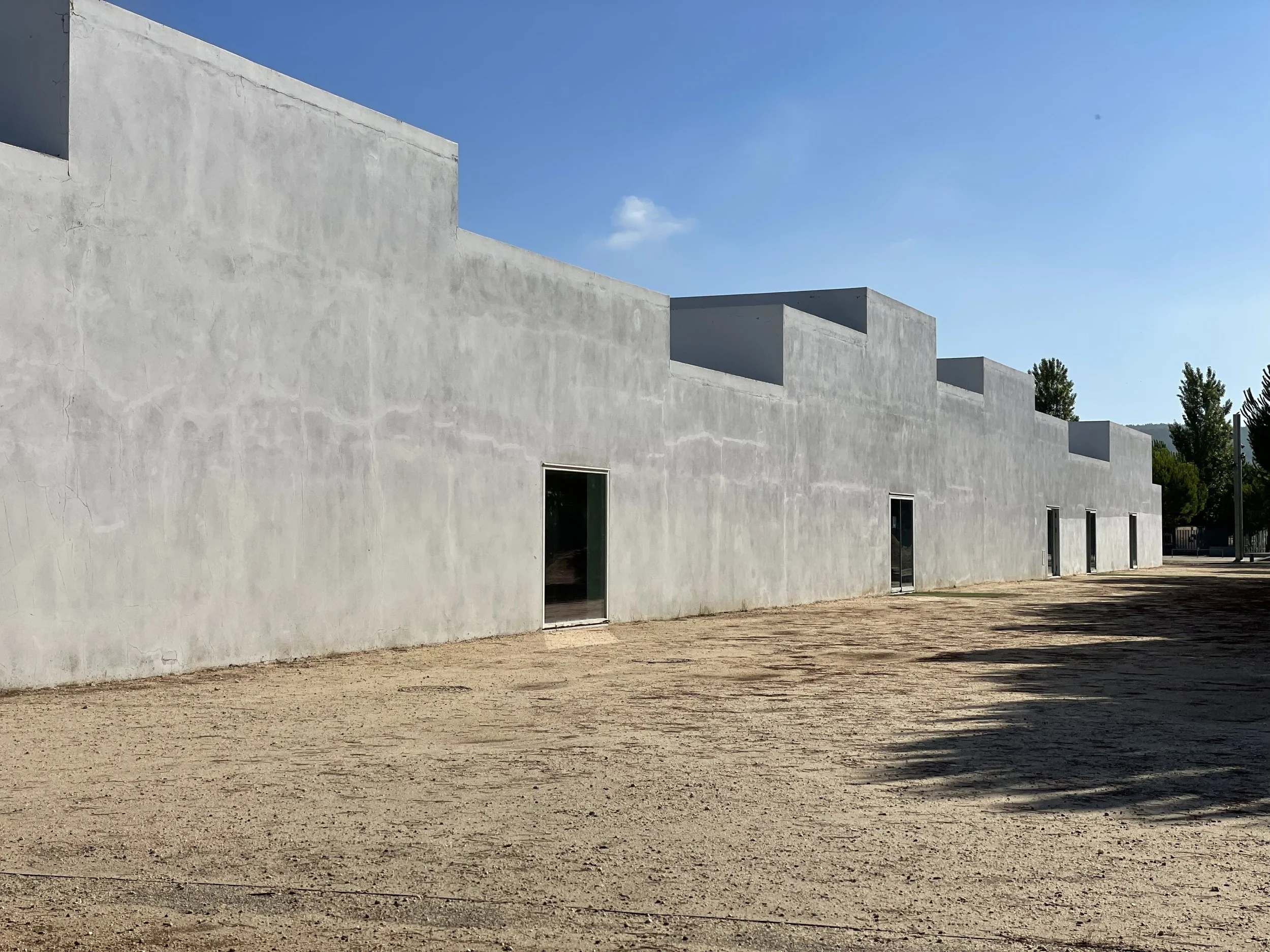 A film and photo location in Portugal from walnut films, A modern, minimalistic white building with flat roofs and rectangular openings, set against a clear blue sky with some trees visible in the background, and a dirt ground in the foreground.