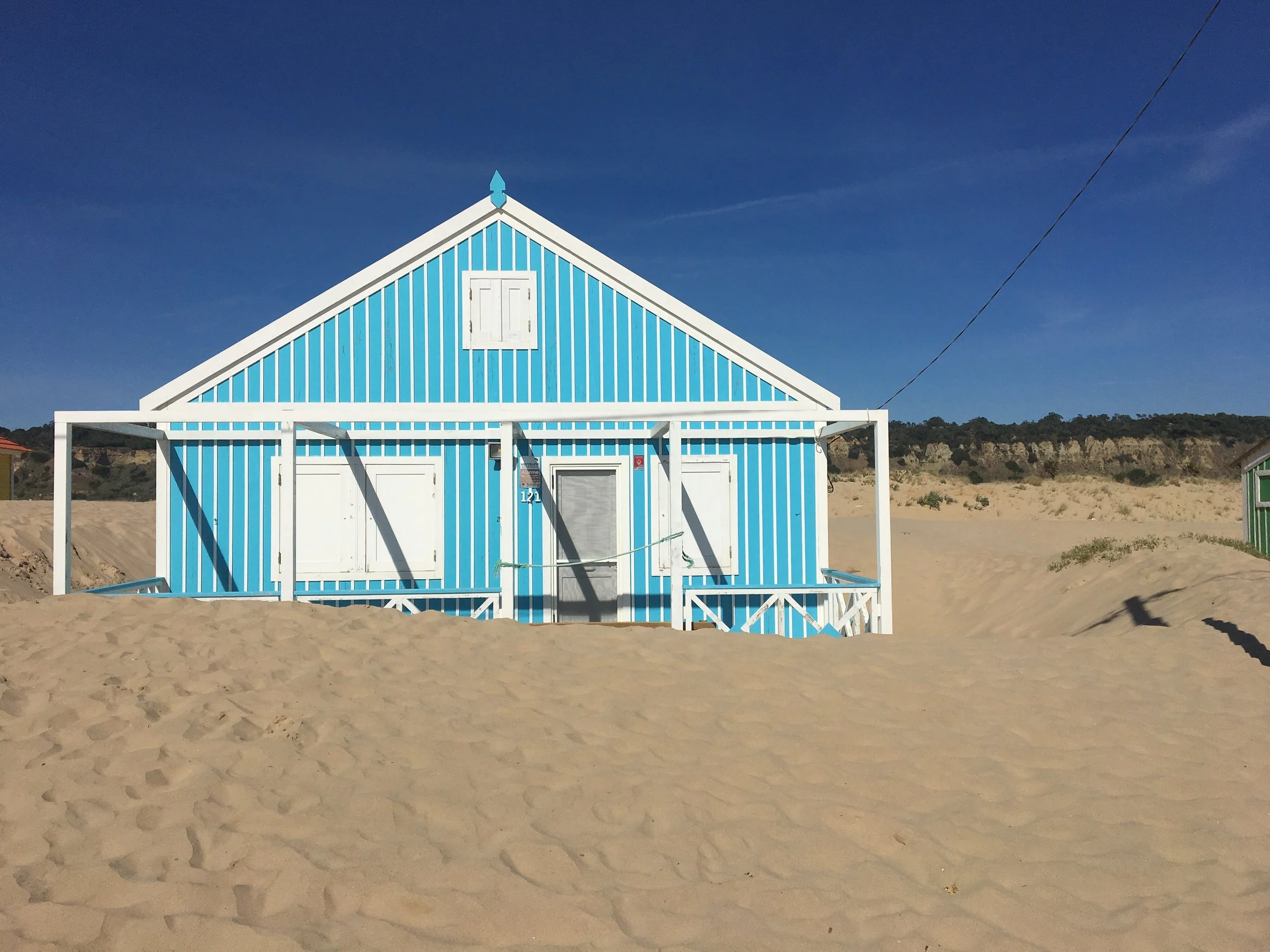 A film and photo location in Portugal from walnut films, A blue and white striped beach house with a small white window at the top and white doors at the bottom, situated on sandy terrain under a clear blue sky.