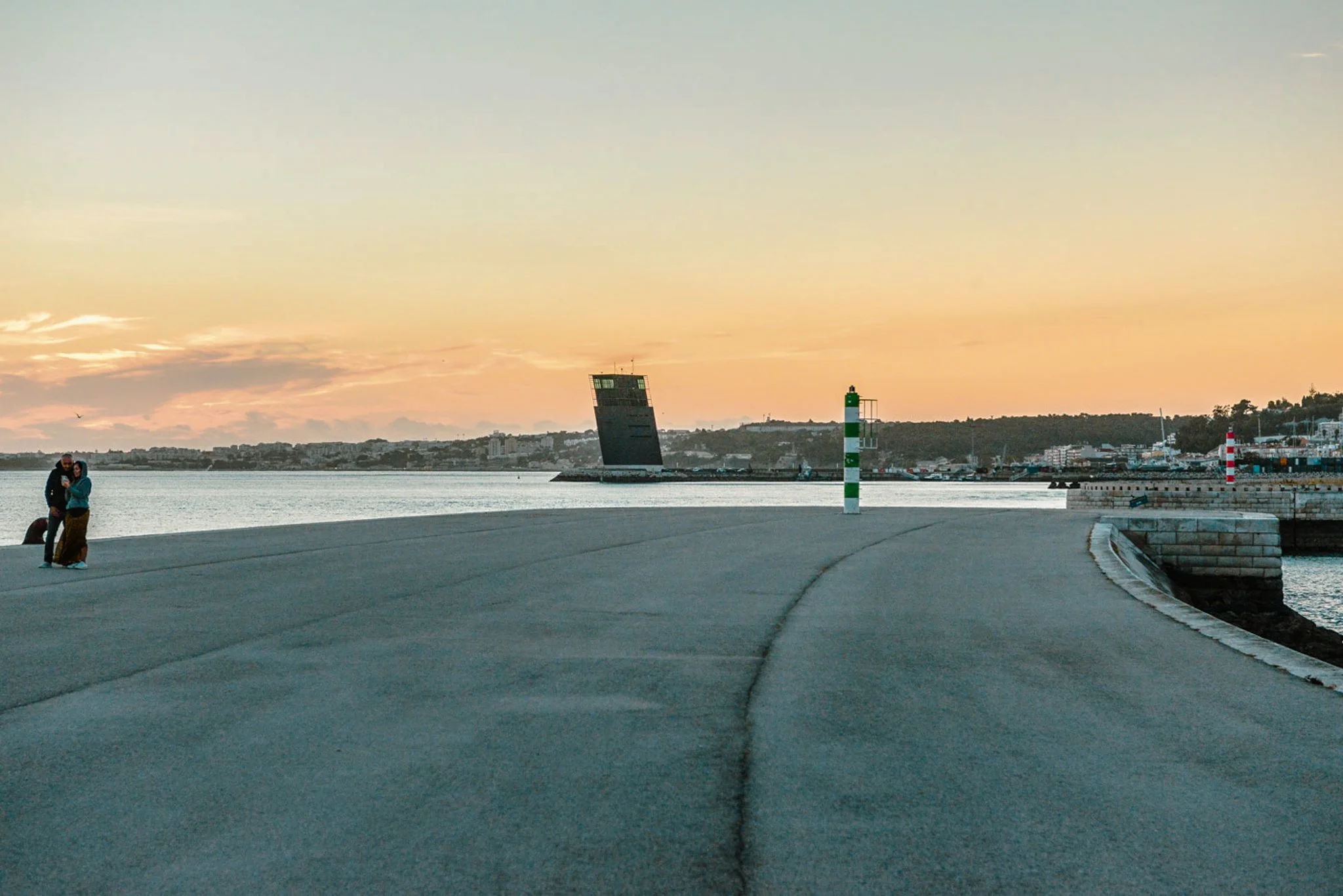 A film and photo location in Portugal from walnut films, A sunset over a harbor with a tilted control tower and a lighthouse, with two people standing near the water's edge.