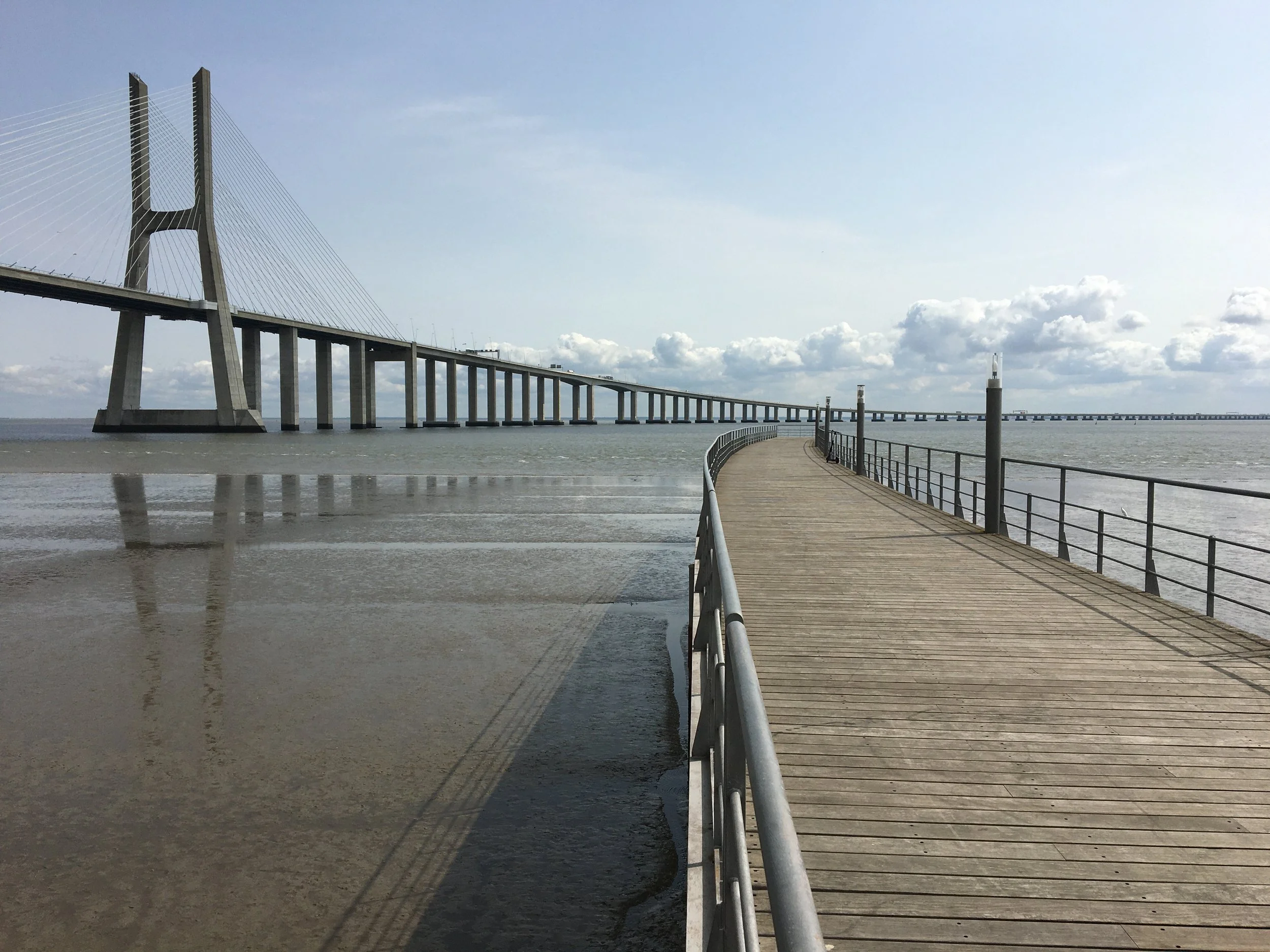 A film and photo location in Portugal from walnut films, A long wooden pier extends over shallow water with the city skyline and clouds in the background. A bridge with tall towers and cables is visible in the distance.