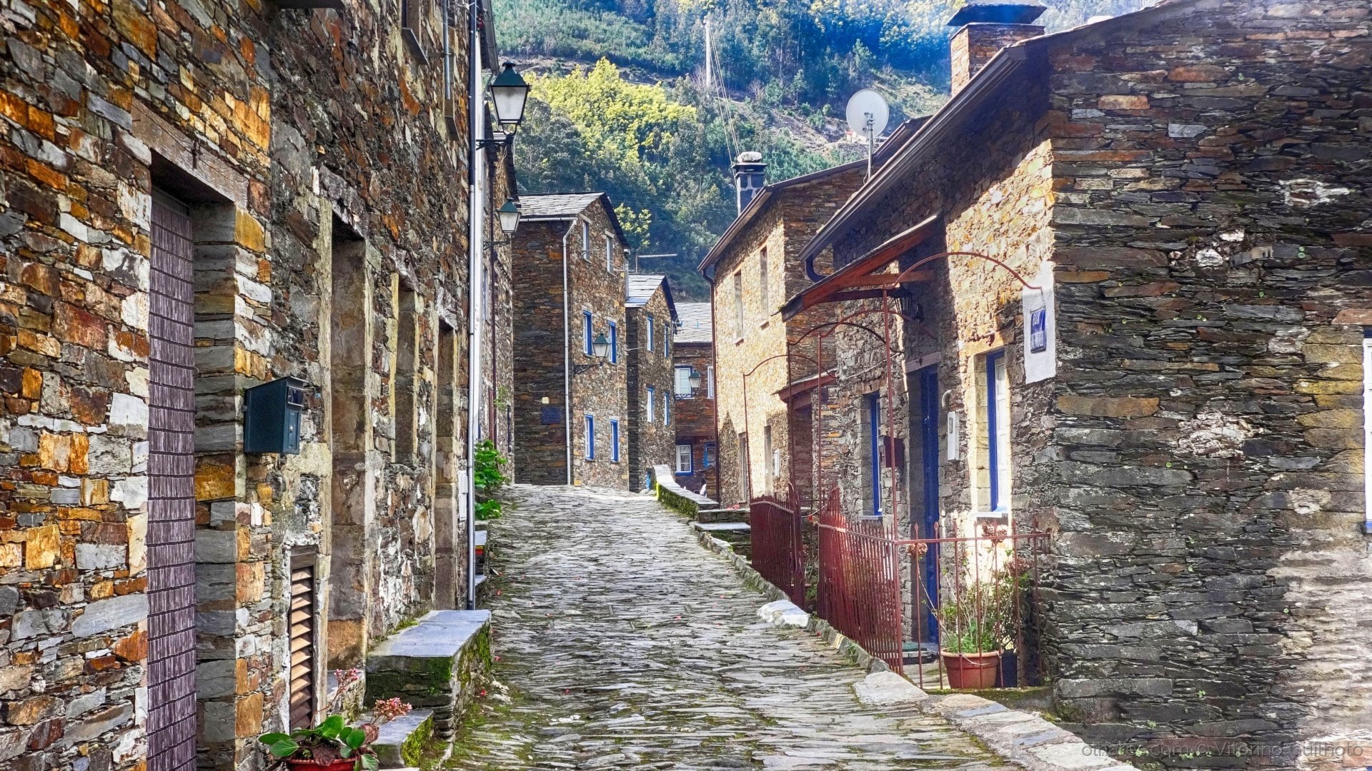 A film and photo location in Portugal from walnut films, A narrow cobblestone street lined with old stone buildings with small windows and doors, some with iron fences, in a mountain village with green hills in the background and clear blue sky.