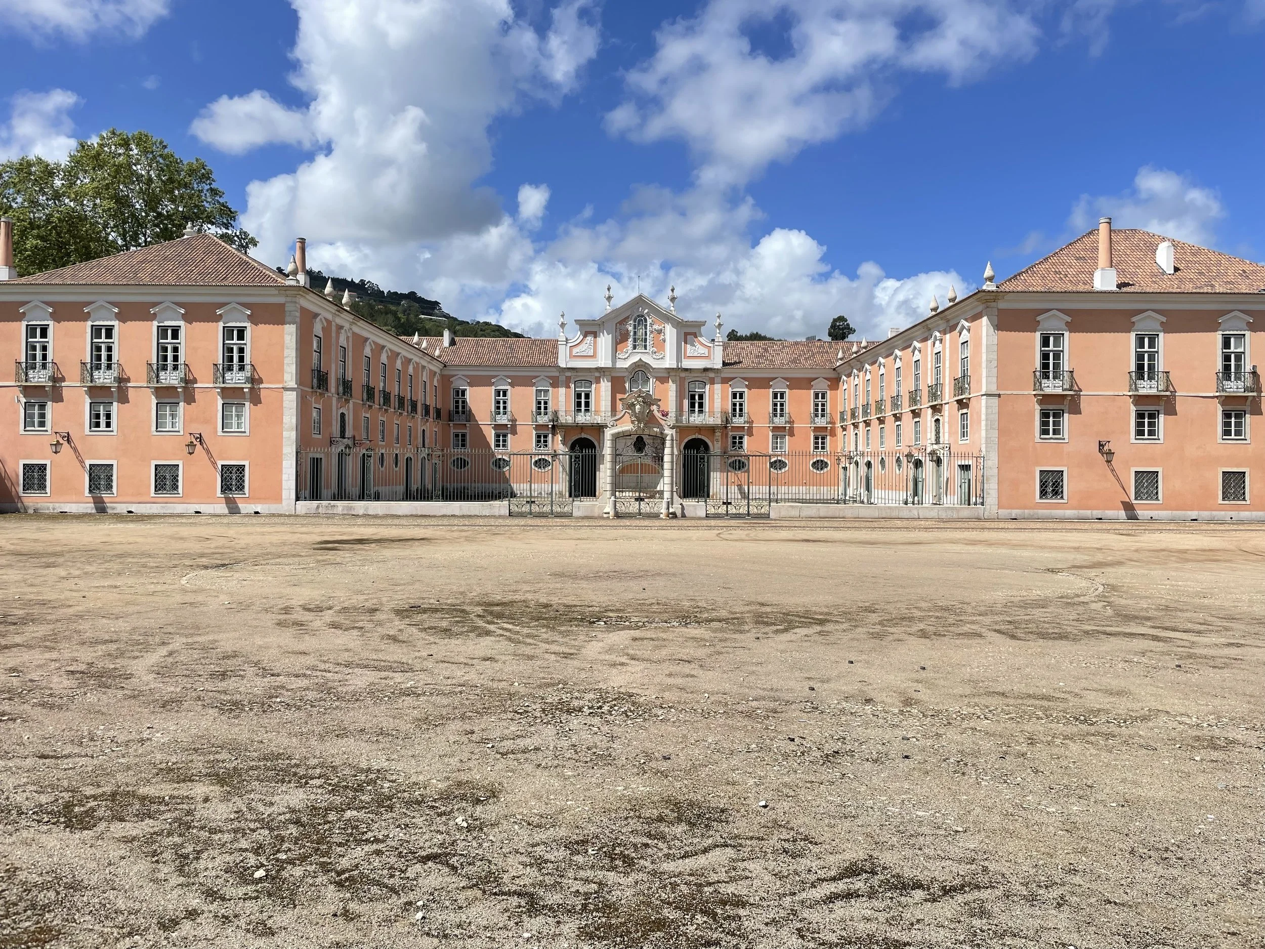 A film and photo location in Portugal from walnut films, A large, historic pink building with white accents and multiple windows, set against a partly cloudy blue sky with trees in the background, surrounded by a dirt area in the foreground.