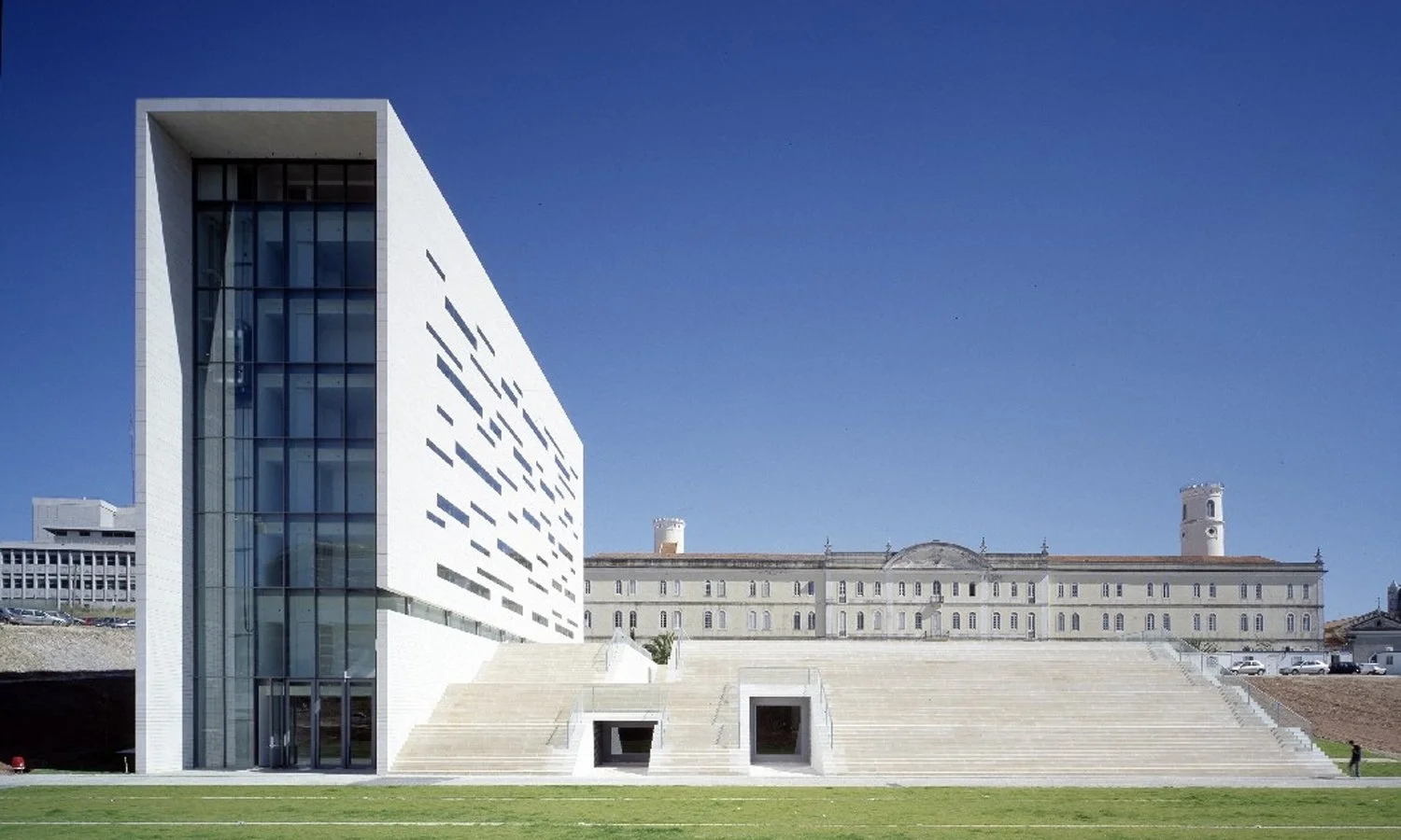 A film and photo location in Portugal from walnut films, Modern white building with large glass windows, an adjacent stone staircase, and a historic building with towers in the background under a clear blue sky.