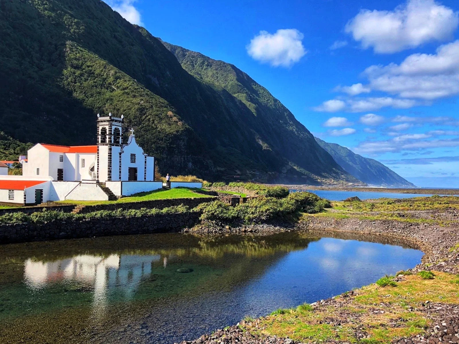 A film and photo location in Portugal from walnut films, A scenic view of a white church with a red-tiled roof, situated on a green grassy area by a body of water. In the background, tall green mountains under a partly cloudy blue sky.